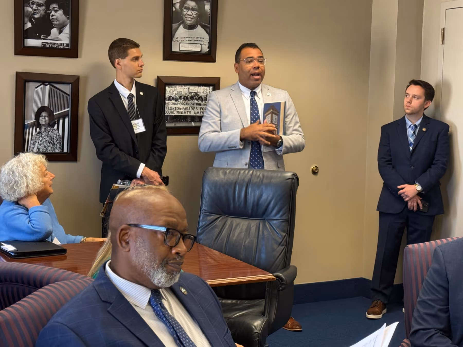 Group of people in a meeting room, one man in a light blazer speaking while holding a photo, others seated or standing listening.