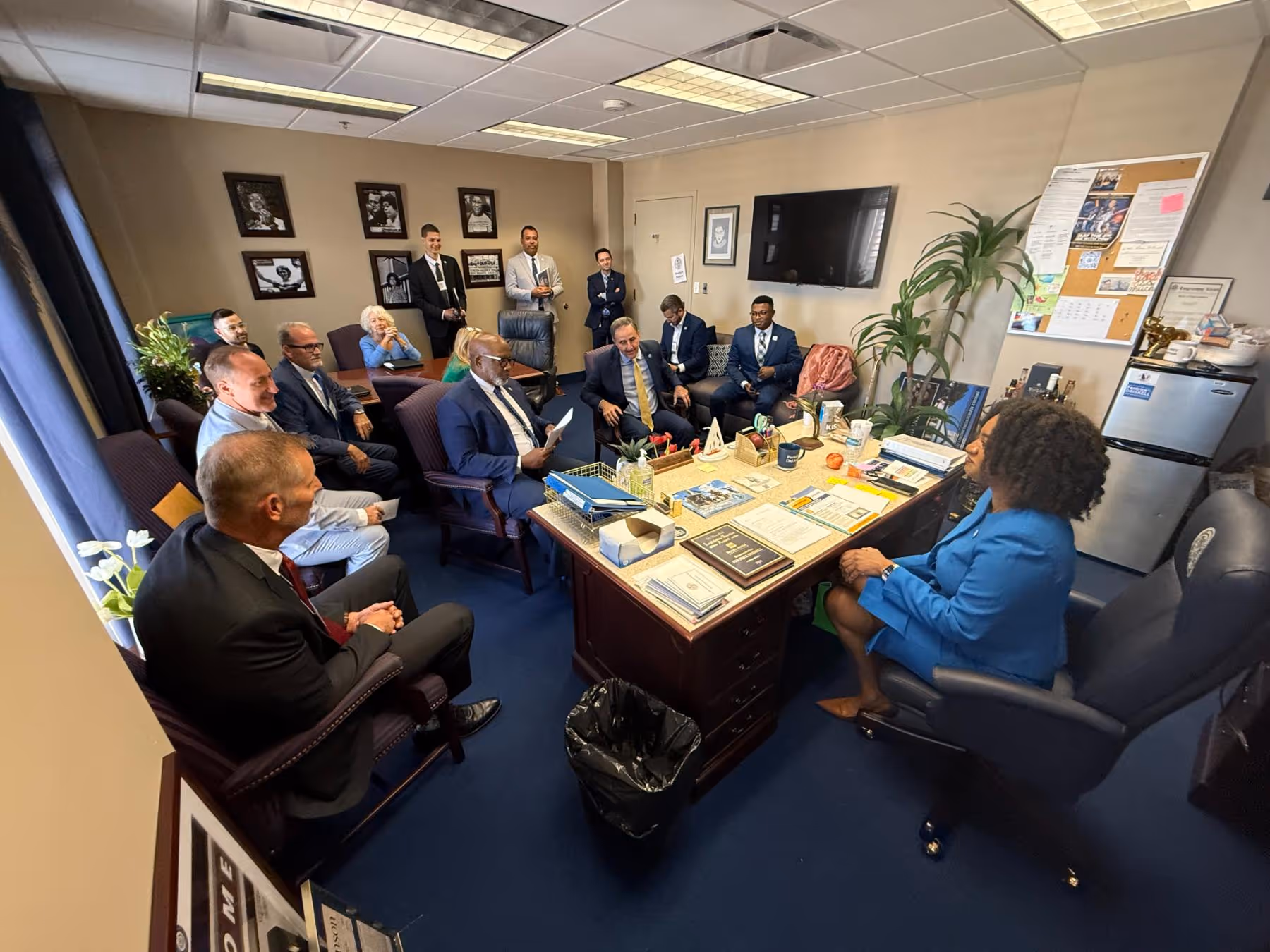 A group of professionally dressed people seated and standing in a well-lit office around a desk covered with papers and office supplies.