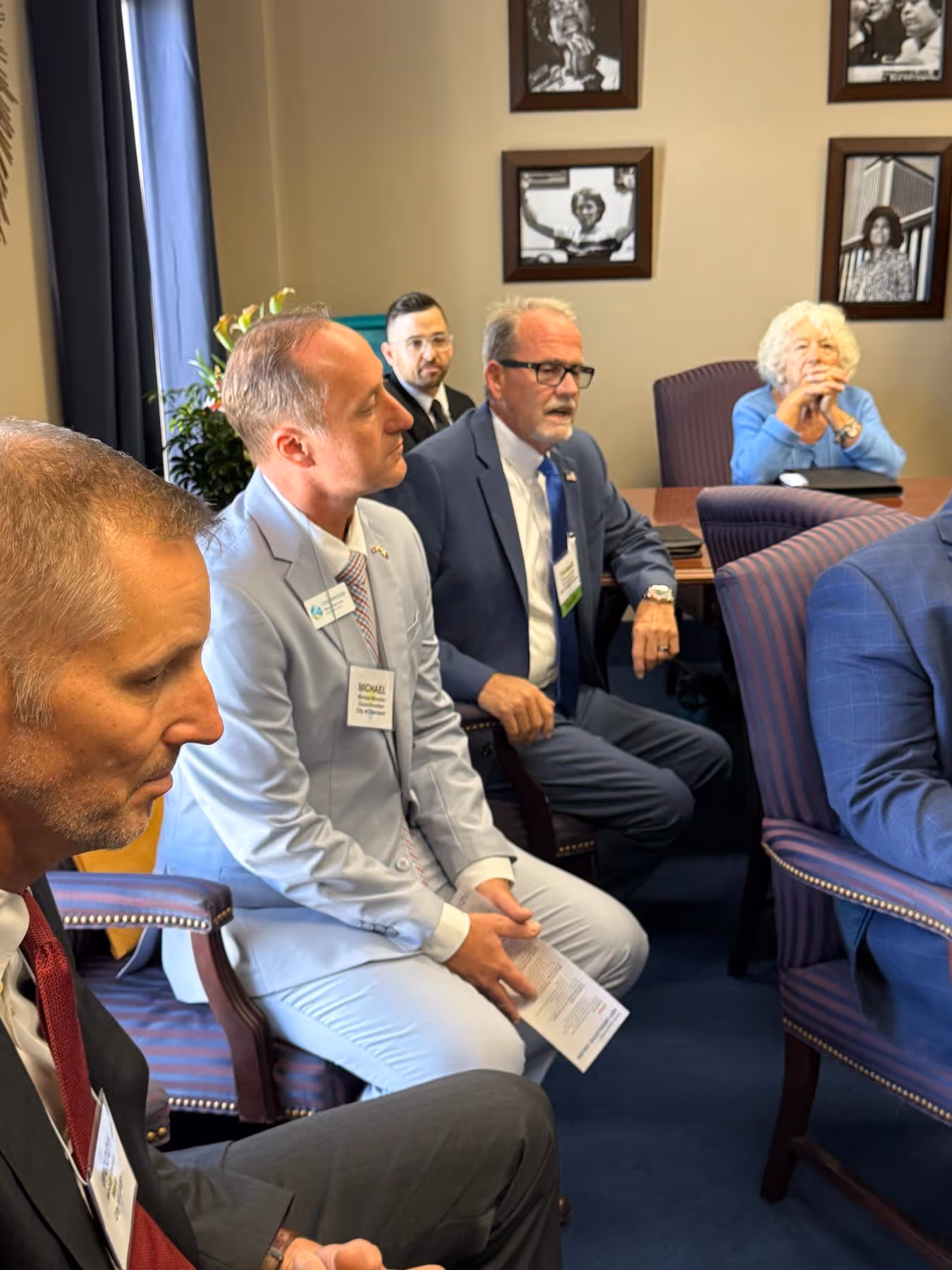 Group of diverse people in business attire seated in a meeting room with framed black-and-white photos on the wall.