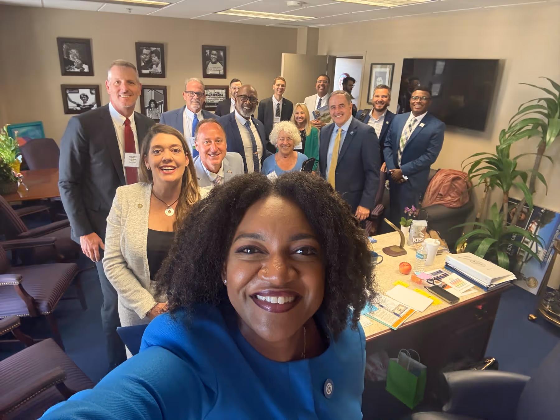 Group selfie of twelve diverse professionals in business attire smiling inside an office.