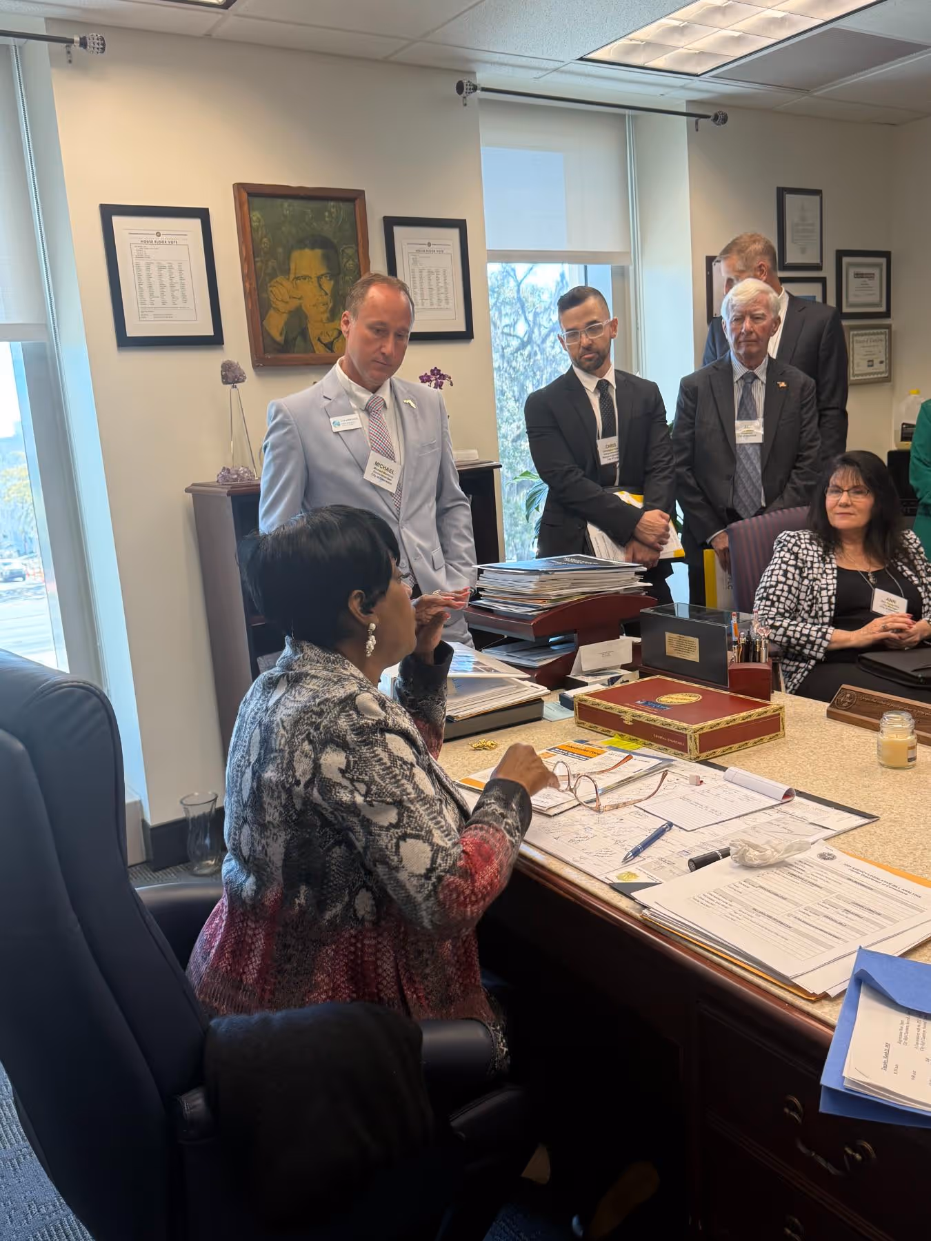 Group of six people in a professional office setting, one woman seated at a desk and five others standing or sitting nearby engaged in discussion.