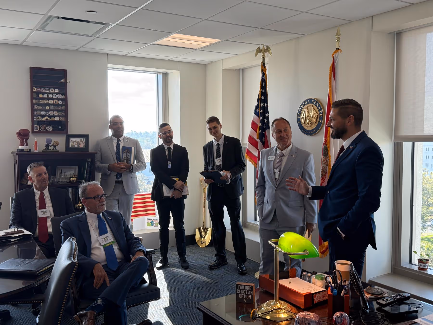 Group of seven men in business attire conversing in an office with Florida Senate emblem, American and Florida state flags, and office desk with green lamp.