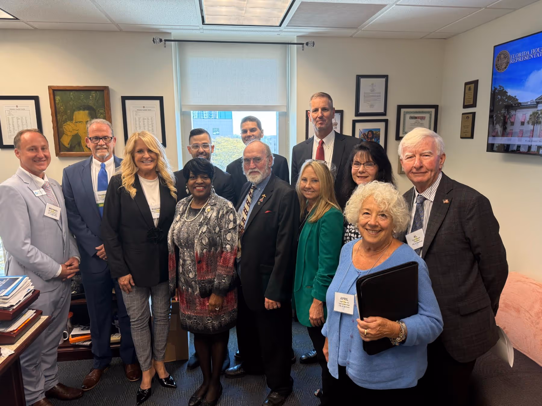 Group of twelve professionally dressed people standing together in an office room with framed documents and a Florida House of Representatives display.
