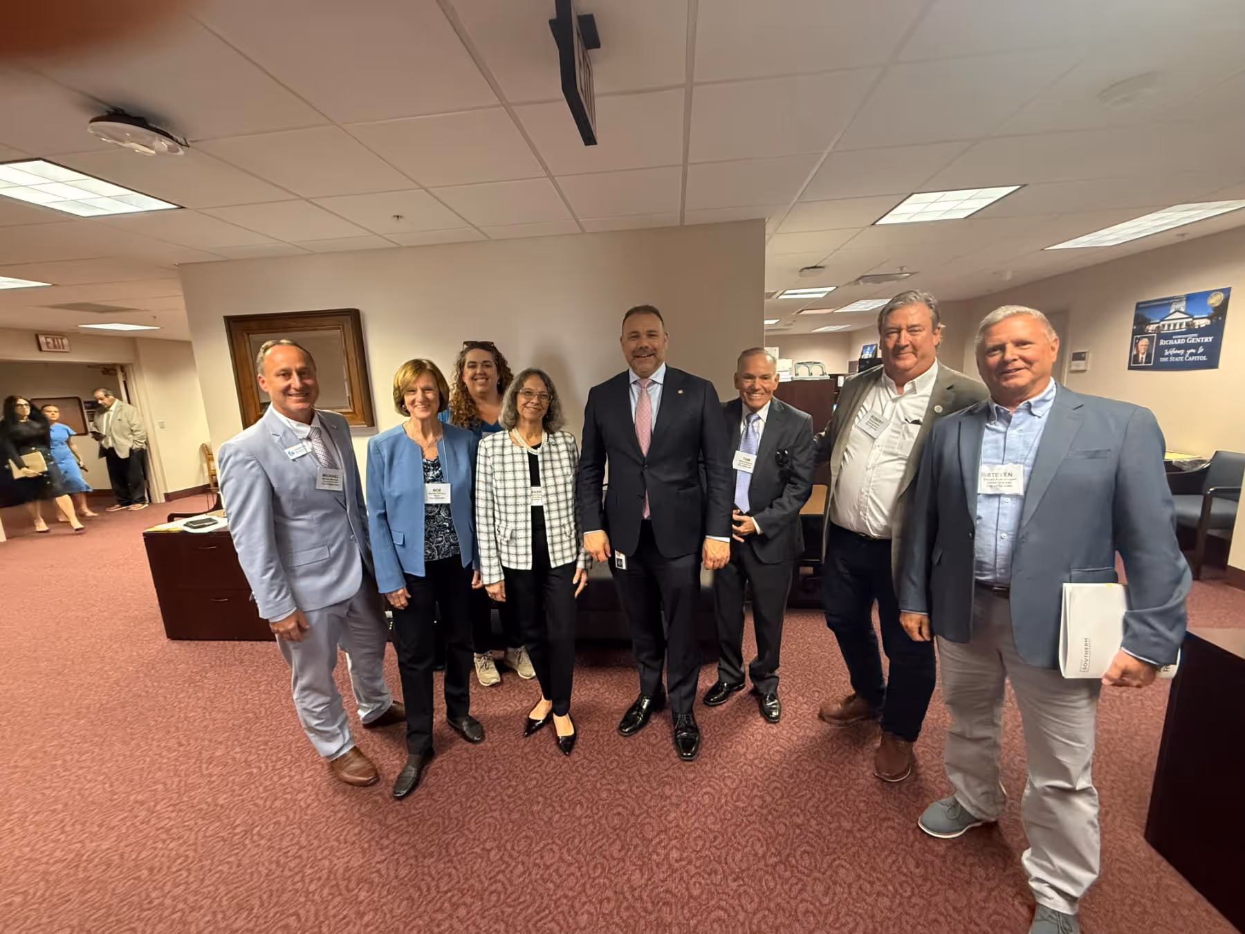 A group of seven people in business attire standing in an office with maroon carpet, smiling at the camera.
