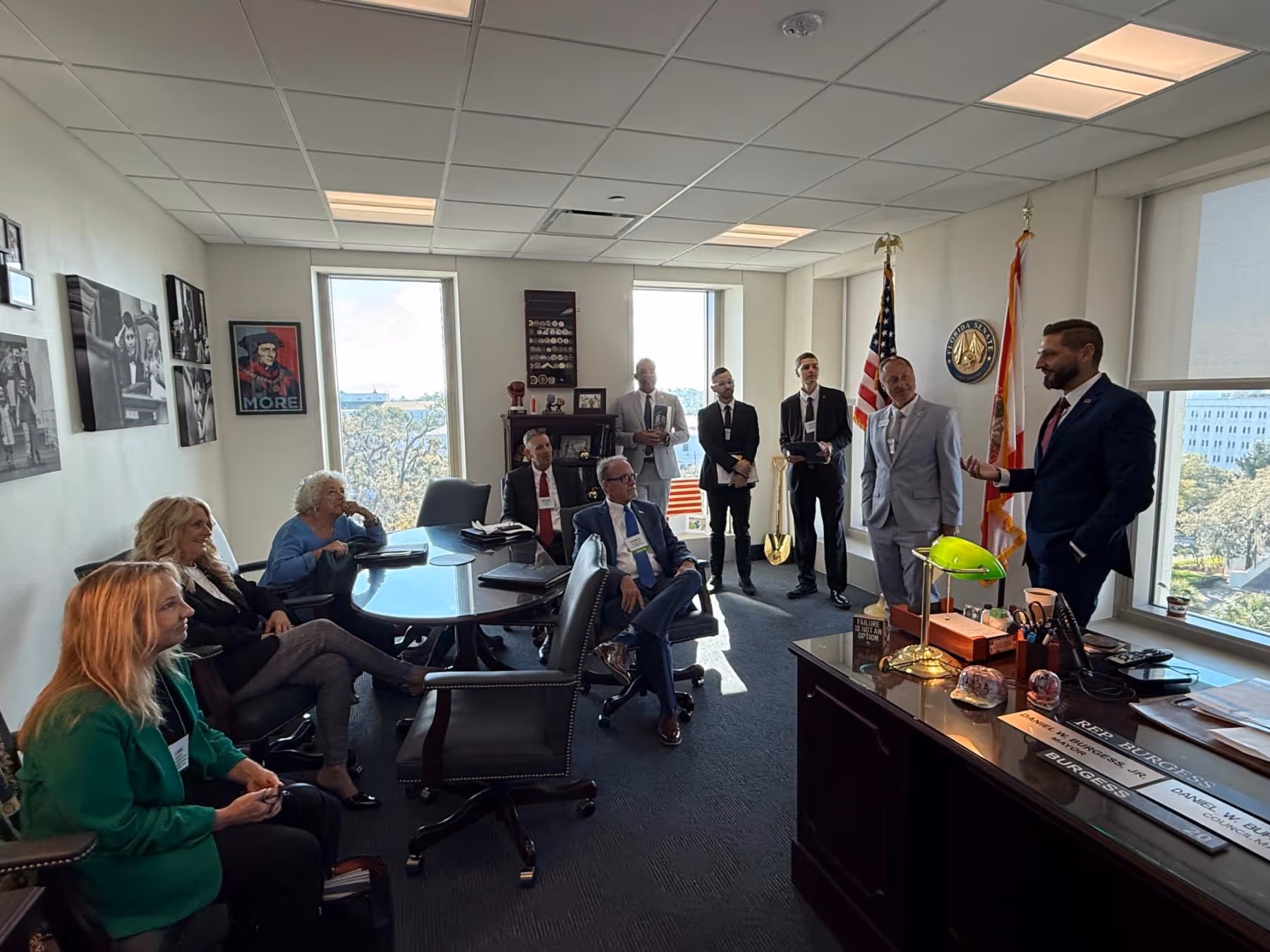 Group of nine people in business attire in an office with flags, a desk, and framed pictures, engaged in a discussion.