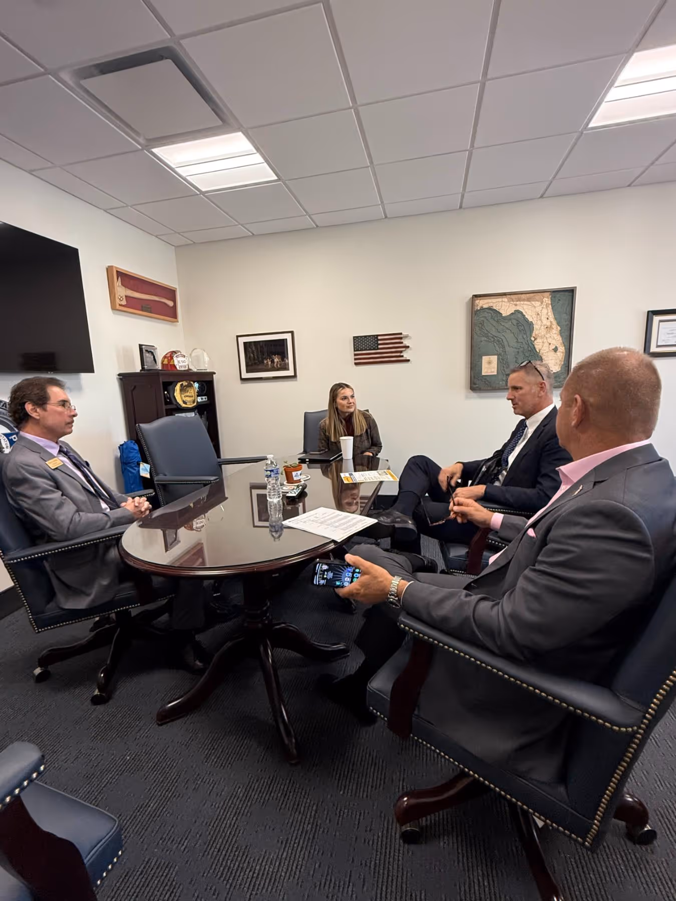 Four people in business attire sitting around a polished oval conference table in a small office engaged in discussion.