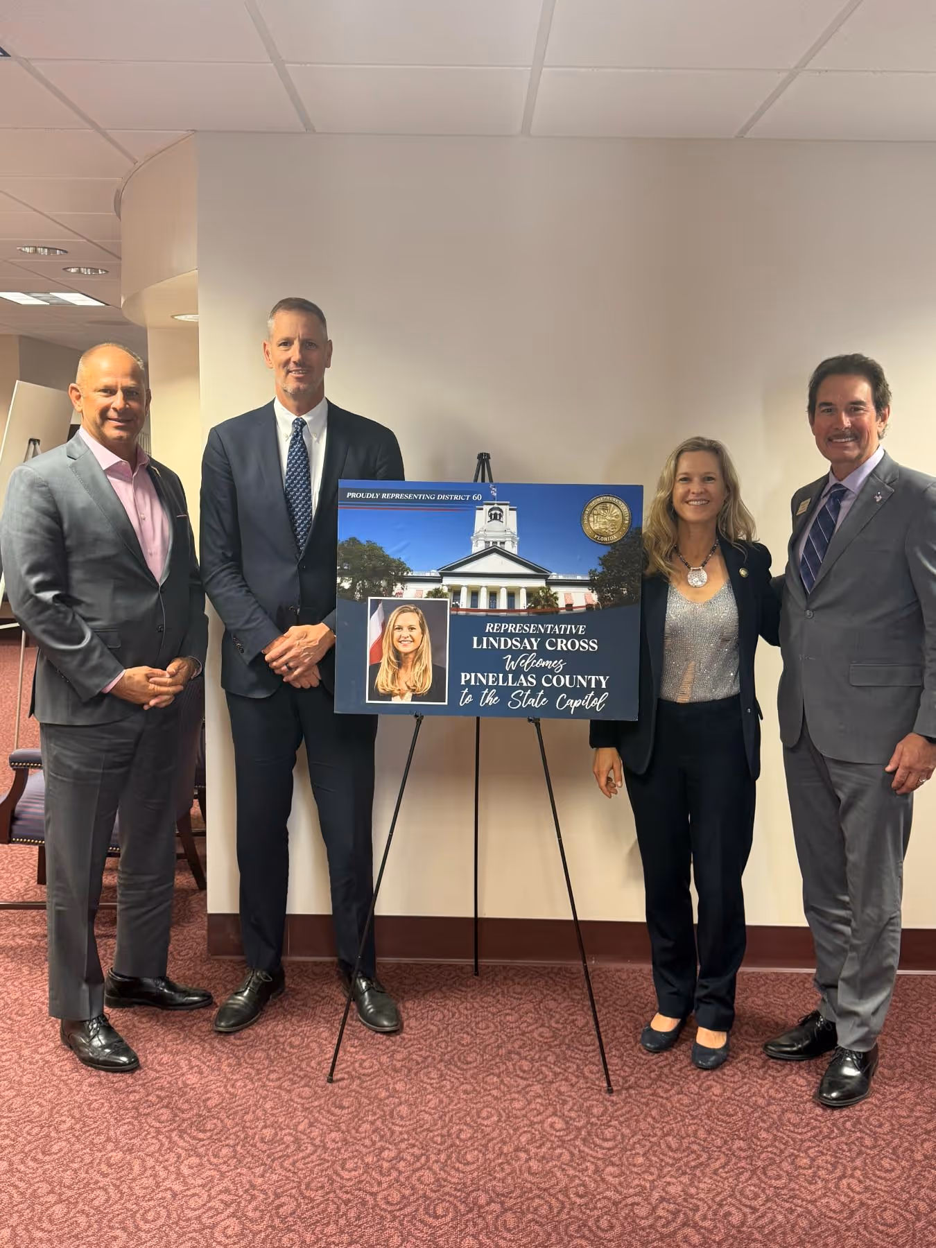 Four people in business attire standing indoors next to a sign that reads 'Representative Lindsay Cross welcomes Pinellas County to the State Capitol.'