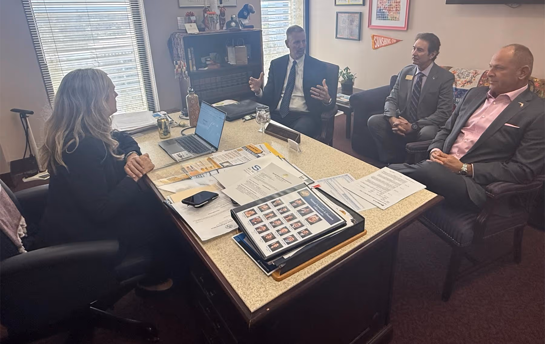 Four adults in business attire having a meeting around a desk cluttered with papers, a laptop, and a binder with photos.