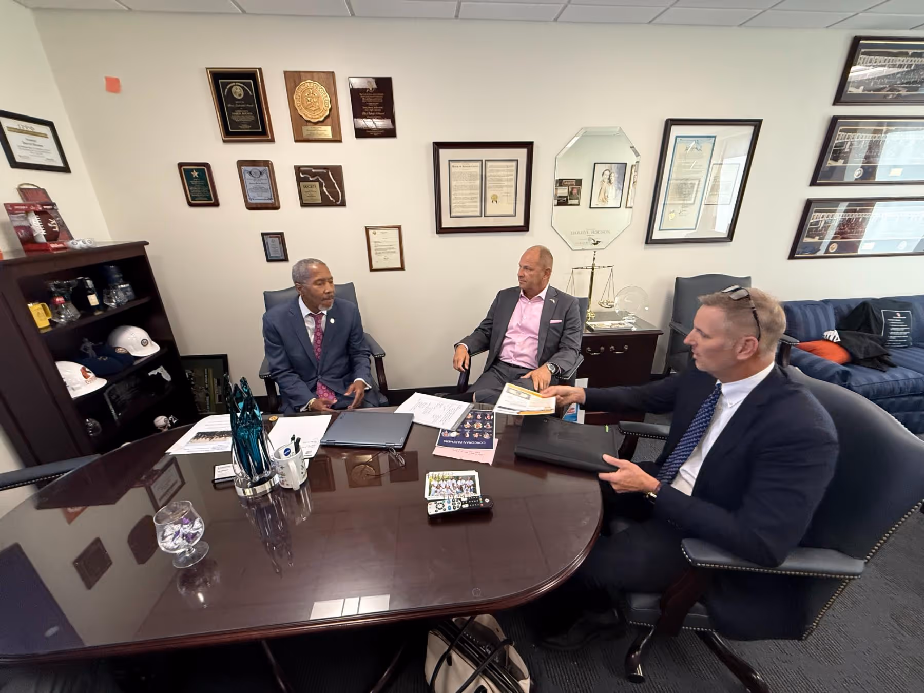 Three men in suits having a meeting around a wooden table in an office with plaques and helmets displayed on shelves and walls.