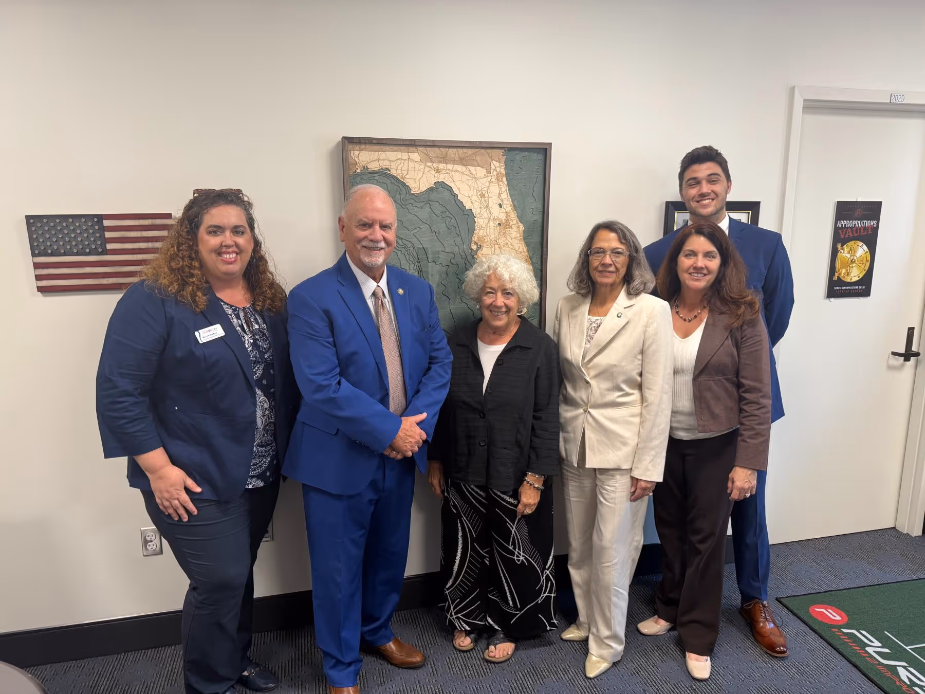 Six people standing together indoors in business and casual attire, with a textured map of Florida on the wall behind them.