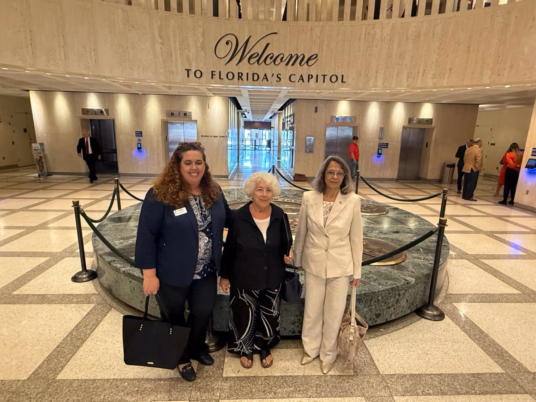 Three women standing inside Florida's Capitol lobby, in front of a circular marble emblem, with 'Welcome to Florida's Capitol' text on the wall above.