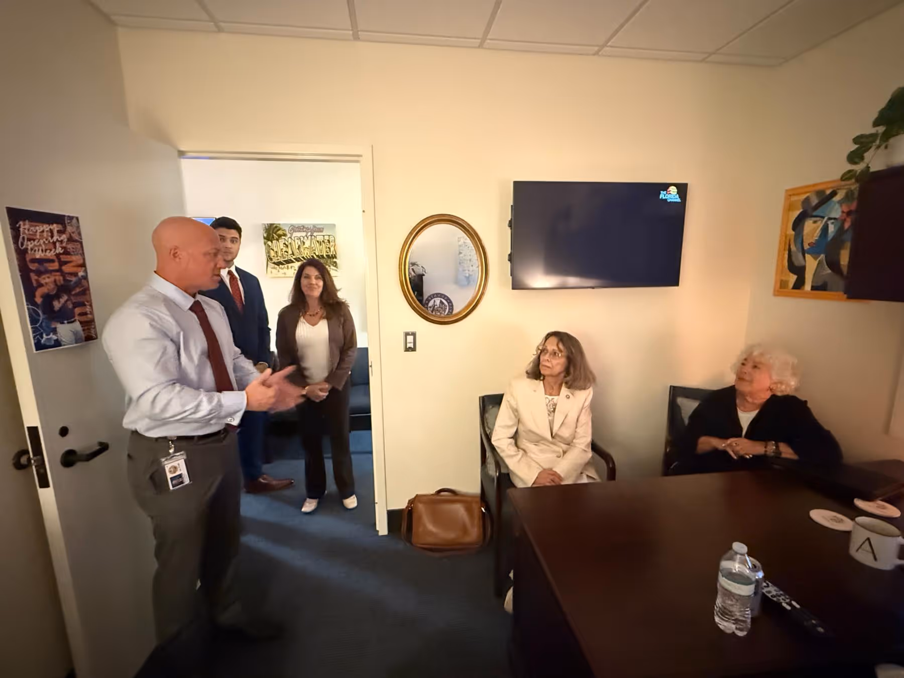 Five people in a small office; one man in a light shirt and tie is speaking while two women sit at a desk and two others stand by the doorway.