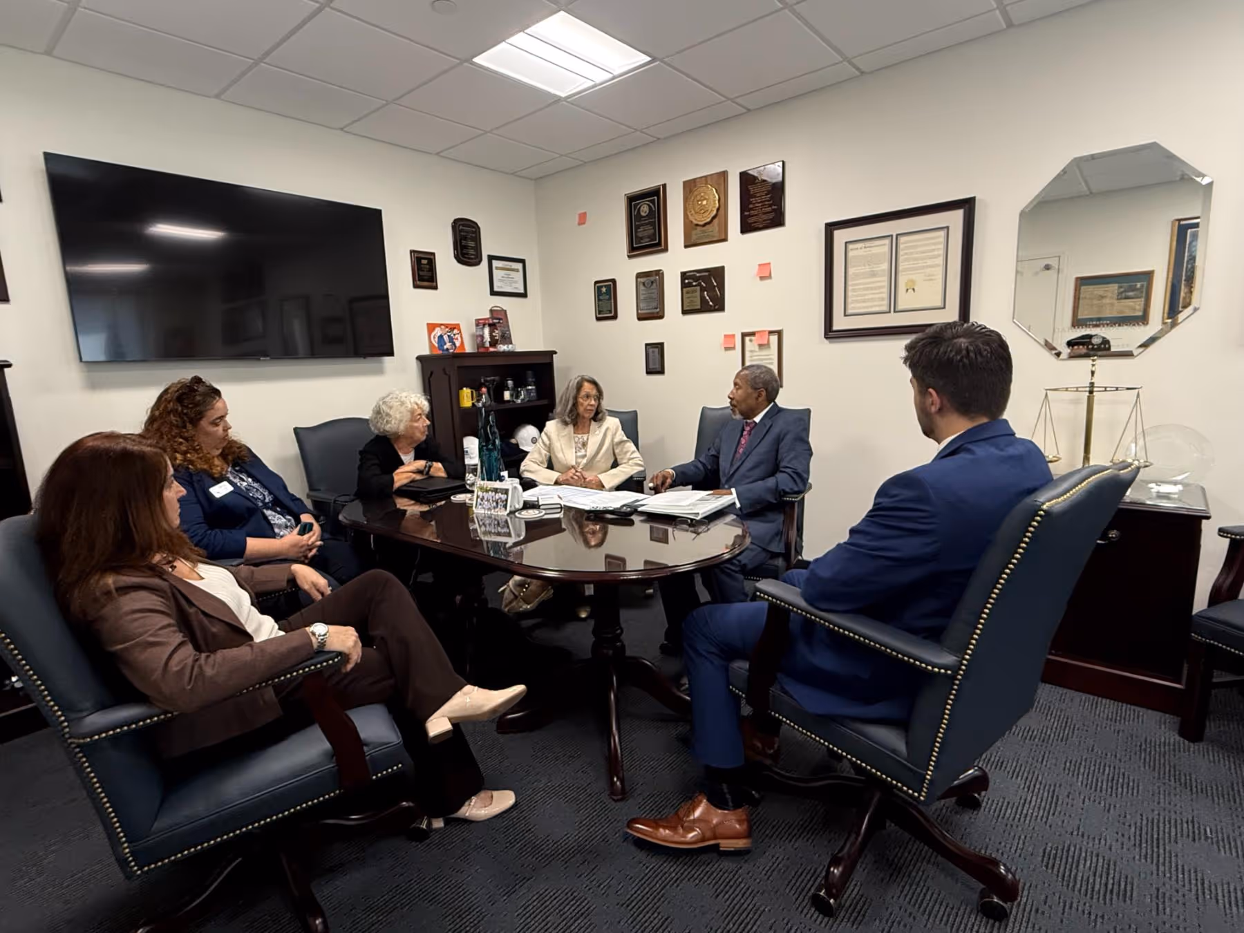 Six people dressed in business attire are seated around a polished wood conference table in an office with plaques on the wall and a large TV screen.