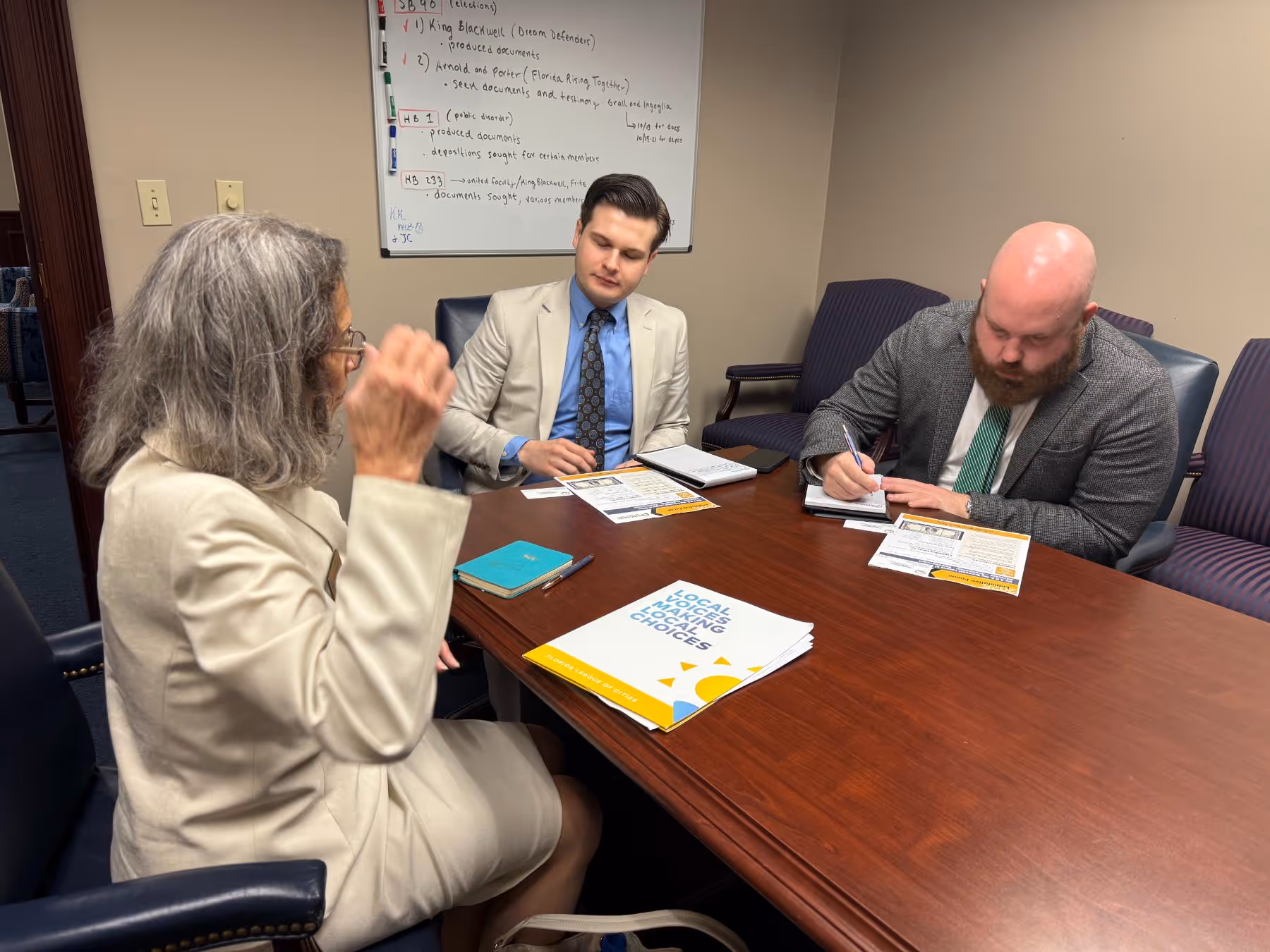 Three professionals in business attire are in a meeting room sitting around a wooden table with papers and a notebook, with a whiteboard displaying handwritten notes in the background.