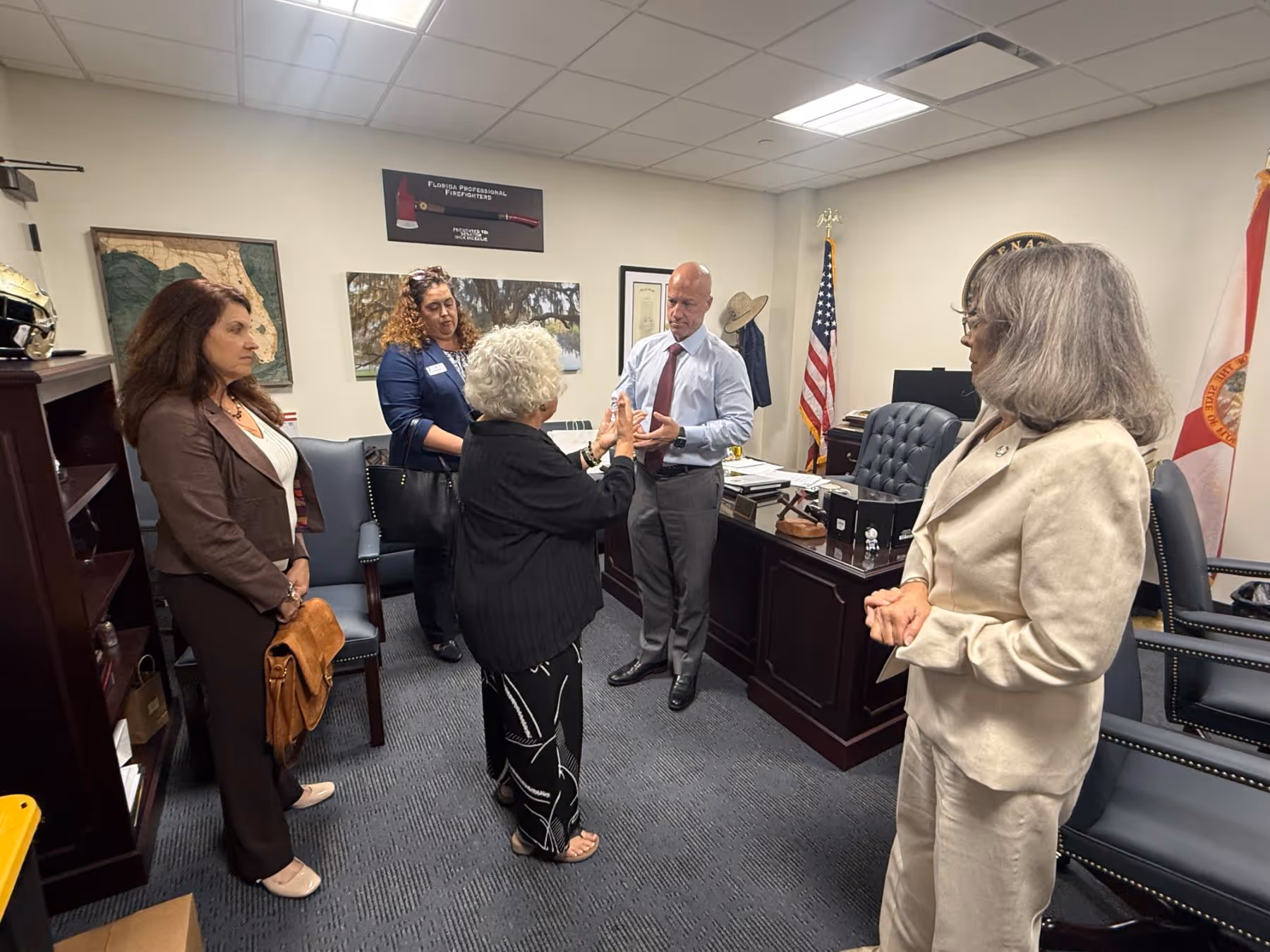 A group of five people having a discussion in an office with American and Florida state flags, maps, and photos on the walls.