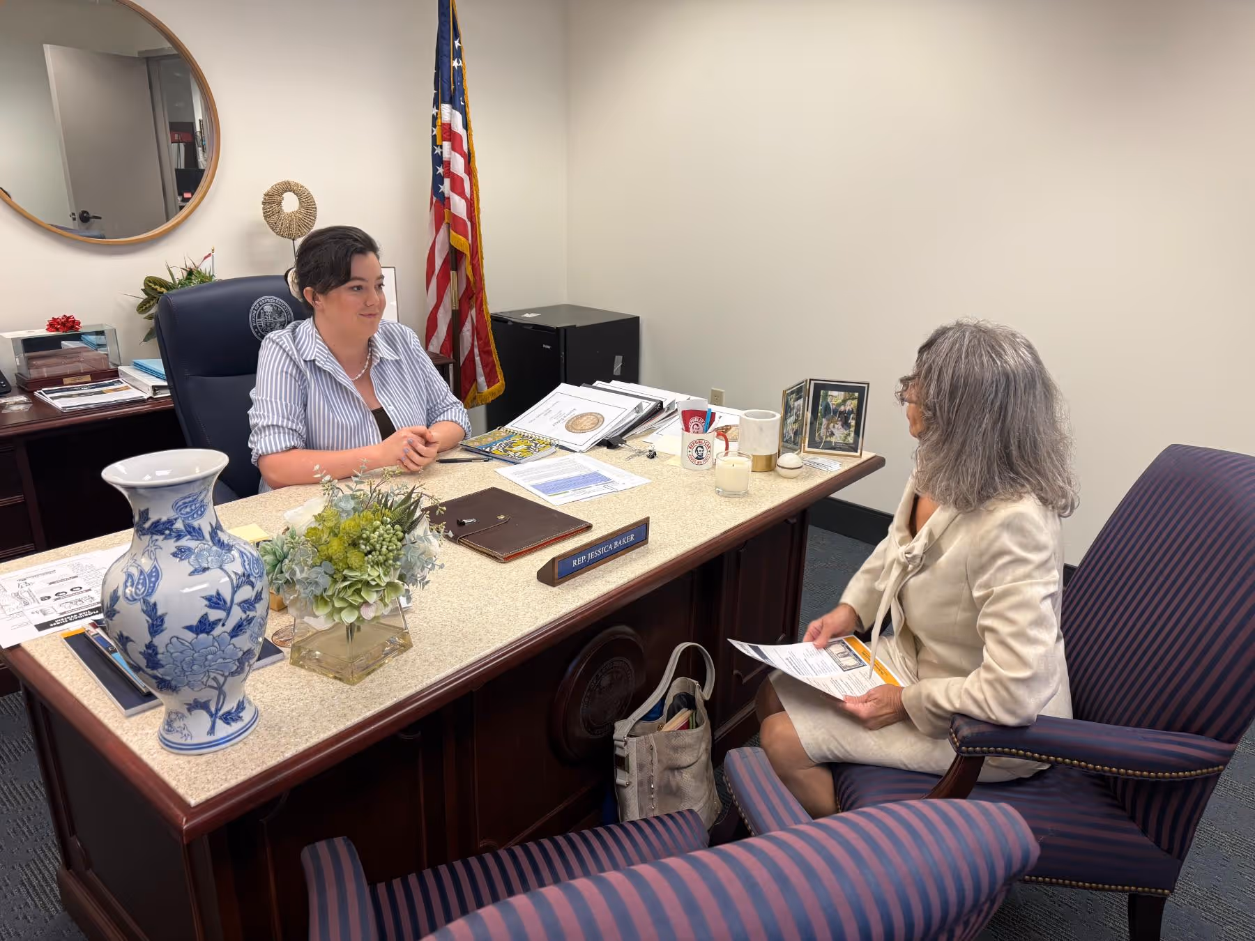 A woman in a striped shirt sits behind a desk labeled Rep Jessica Baker, talking to another woman with gray hair in a cream suit holding papers.