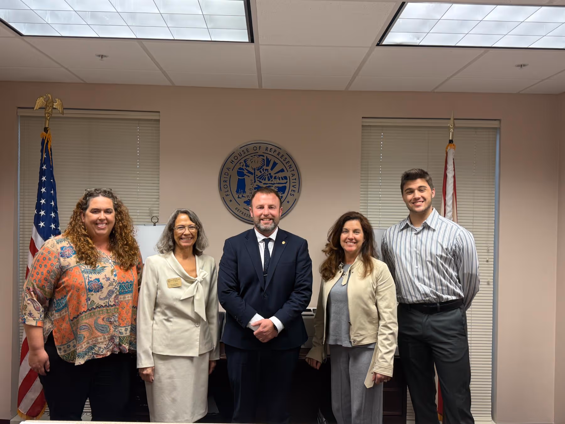 Five people standing and smiling in an office with Florida House of Representatives seal and two flags behind them.