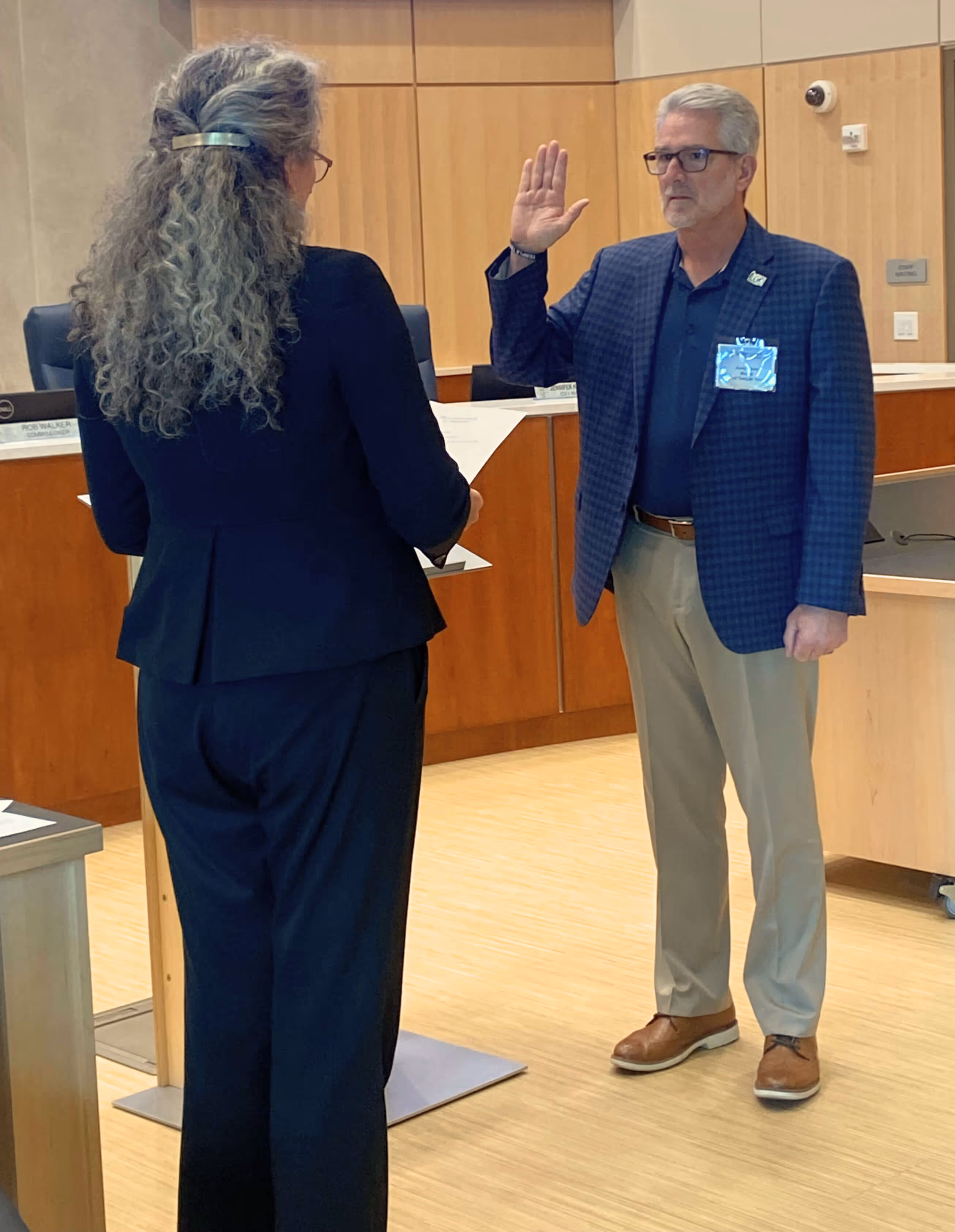 Man in glasses and blazer raising his right hand, possibly taking an oath, facing a woman holding a paper in an indoor formal setting.