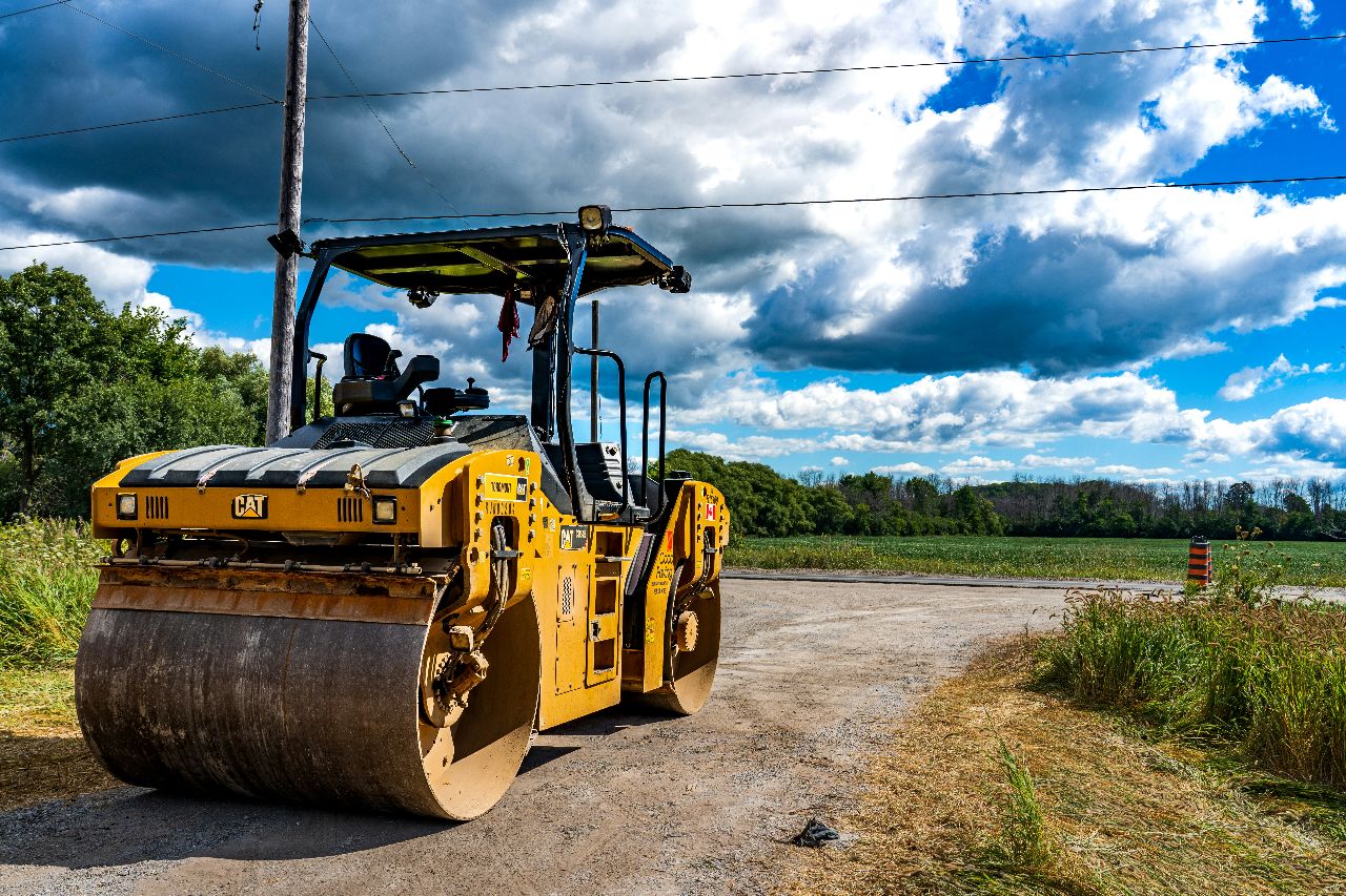 A yellow CAT vibratory road roller sits idle on a rural construction site under a dramatic, cloud-filled blue sky.