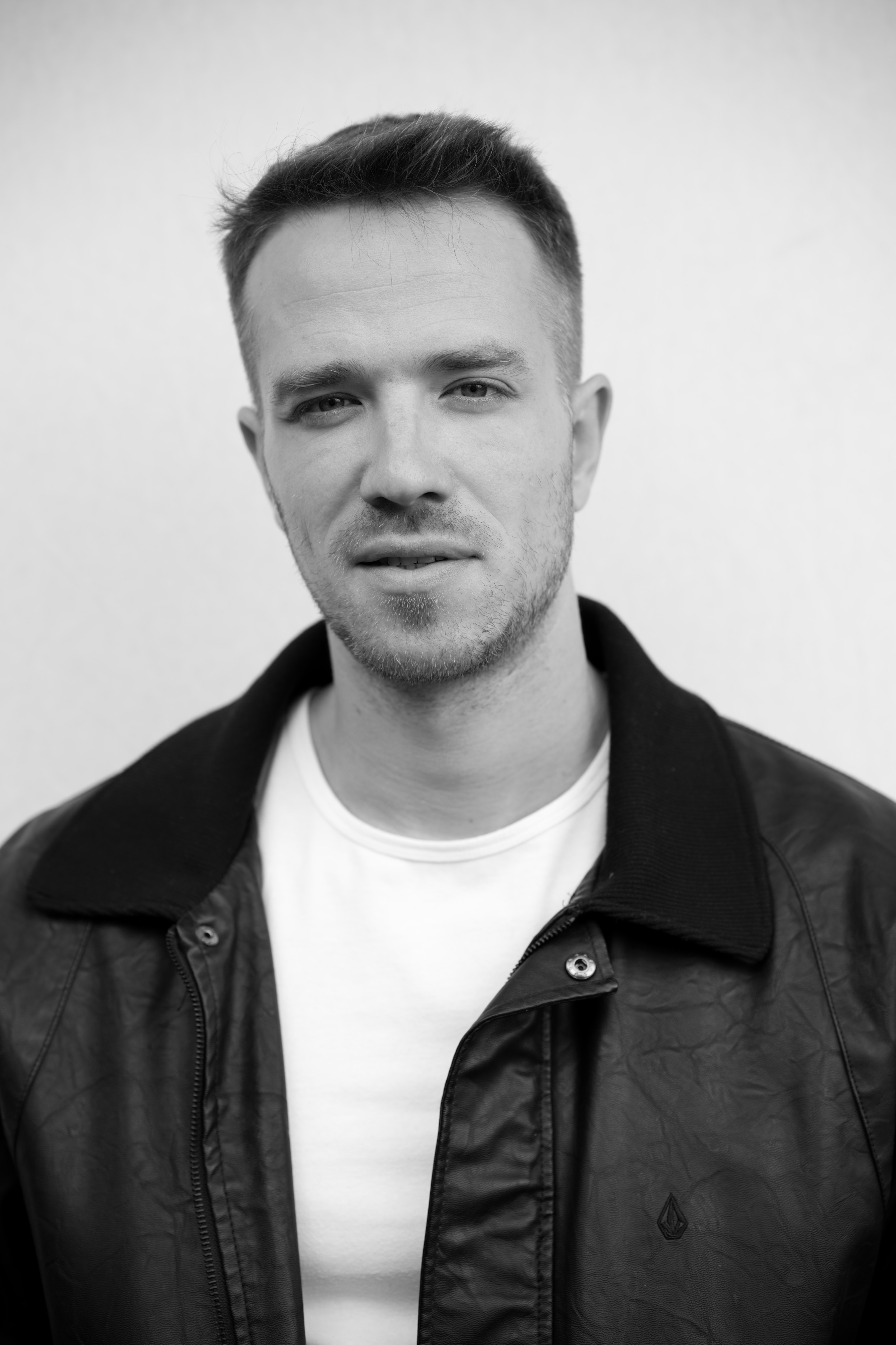 Black-and-white portrait of a man with short hair, light facial stubble, wearing a white shirt and a leather jacket.