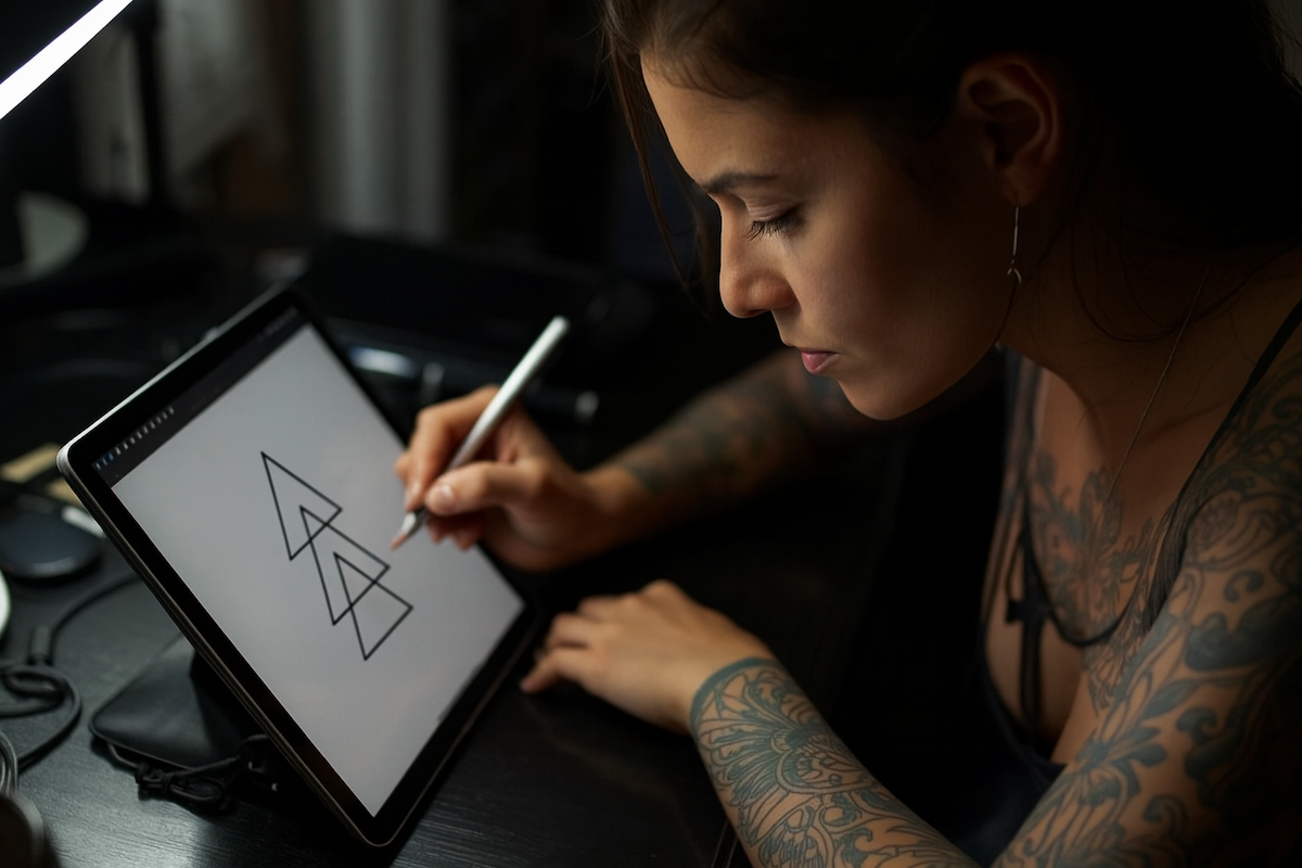 Woman with tattoos focused on drawing overlapping geometric triangles on a tablet with a stylus.