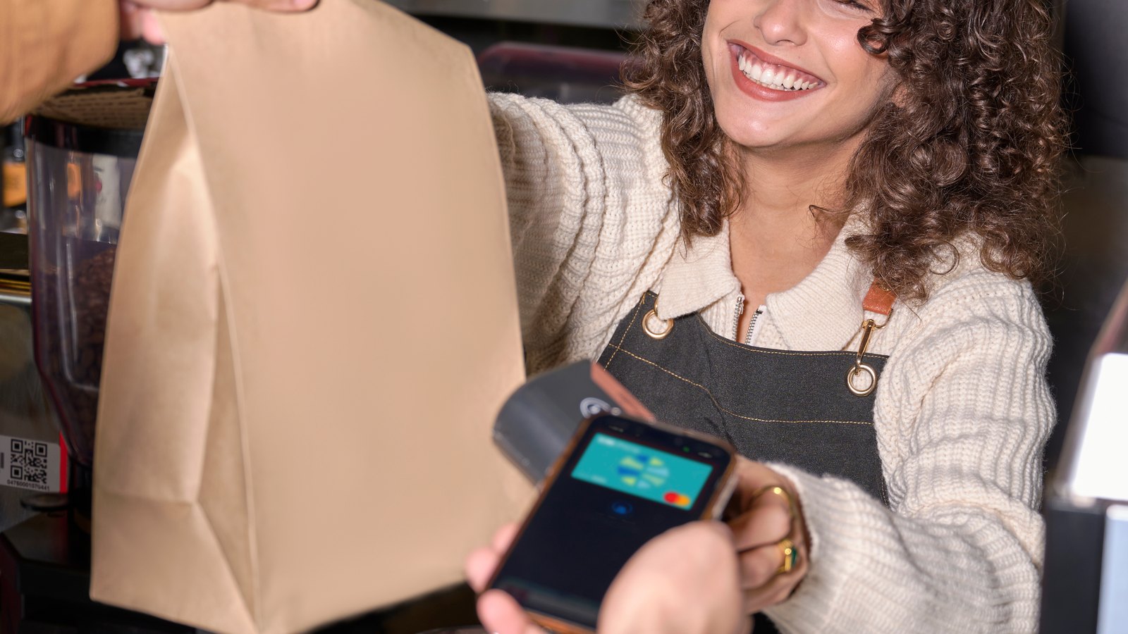 A smiling barista hands a Zeal payment terminal across a cafe counter to a customer, the terminal screen showing a loyalty reward alongside the card payment.