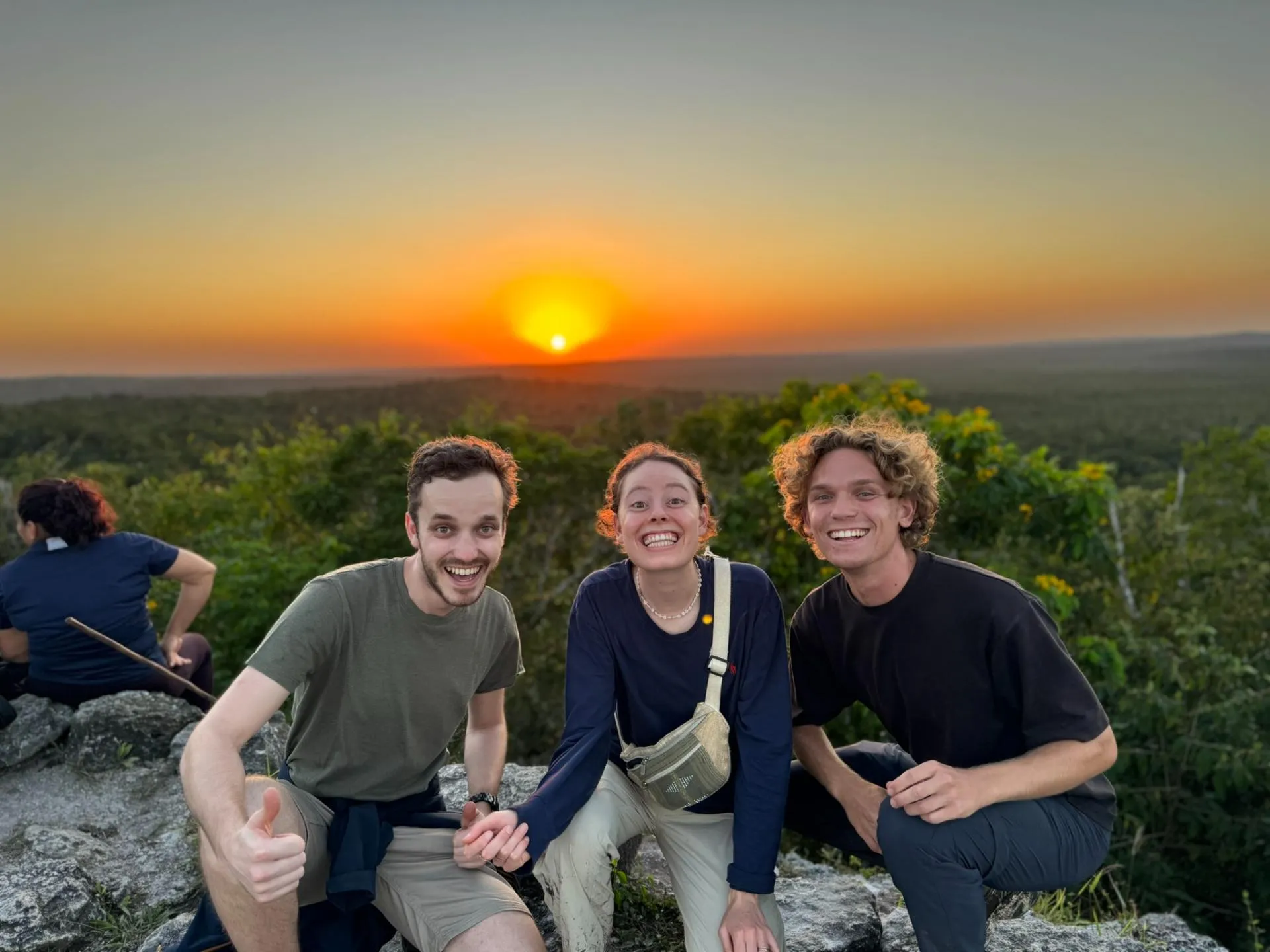 Three friends smiling and sitting on rocks with a forest and a vibrant sunset in the background.