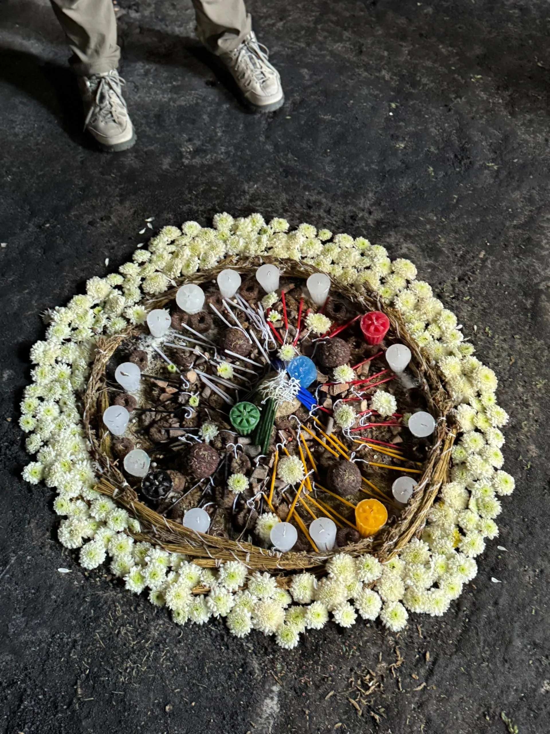 Circular floral arrangement on the ground with white chrysanthemums surrounding colored candles and twigs, with a person wearing beige shoes standing nearby.