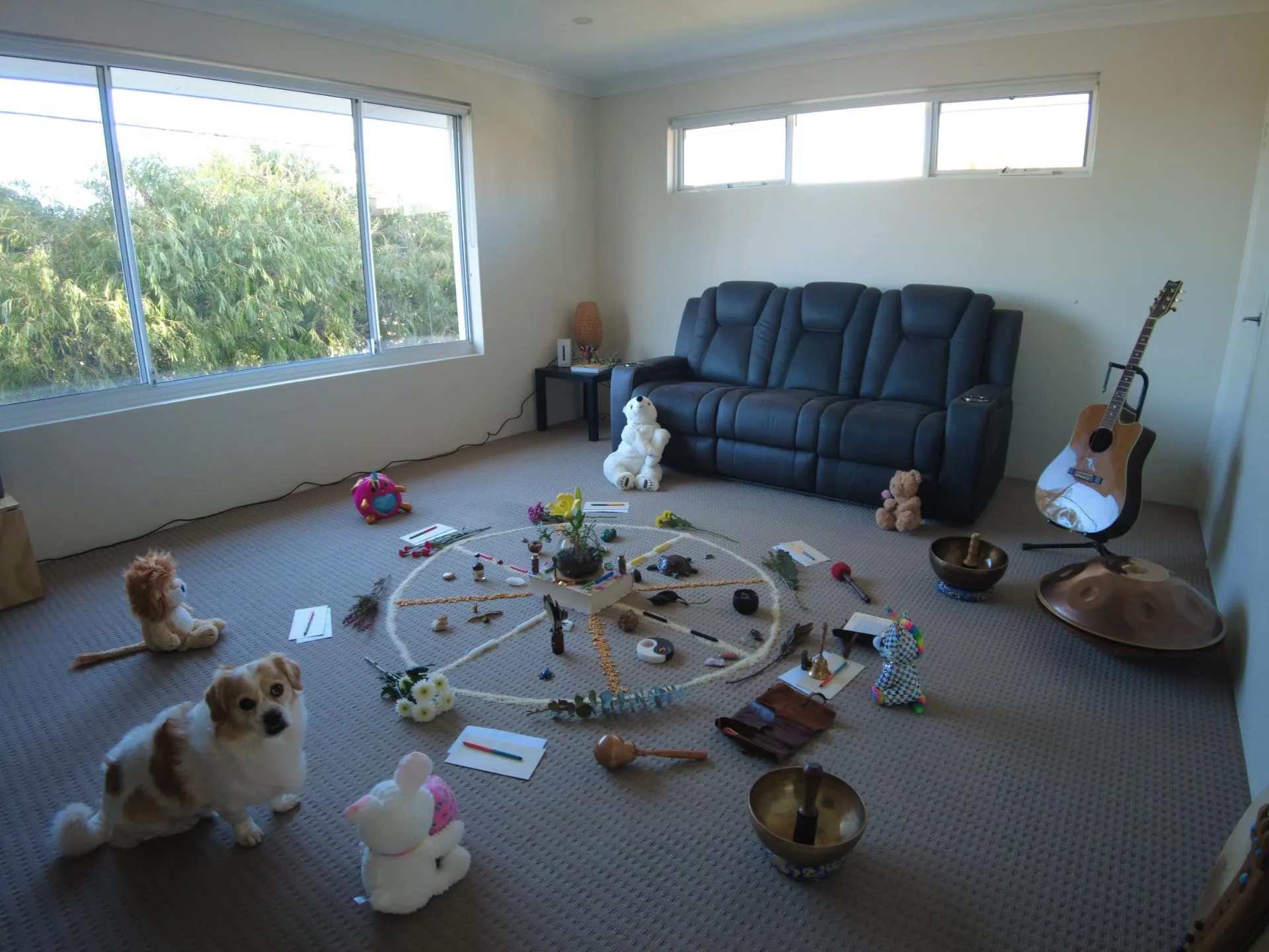 Carpeted room with a dark sofa, a guitar, a handpan, and a circular arrangement of flowers, toys, and small objects on the floor.