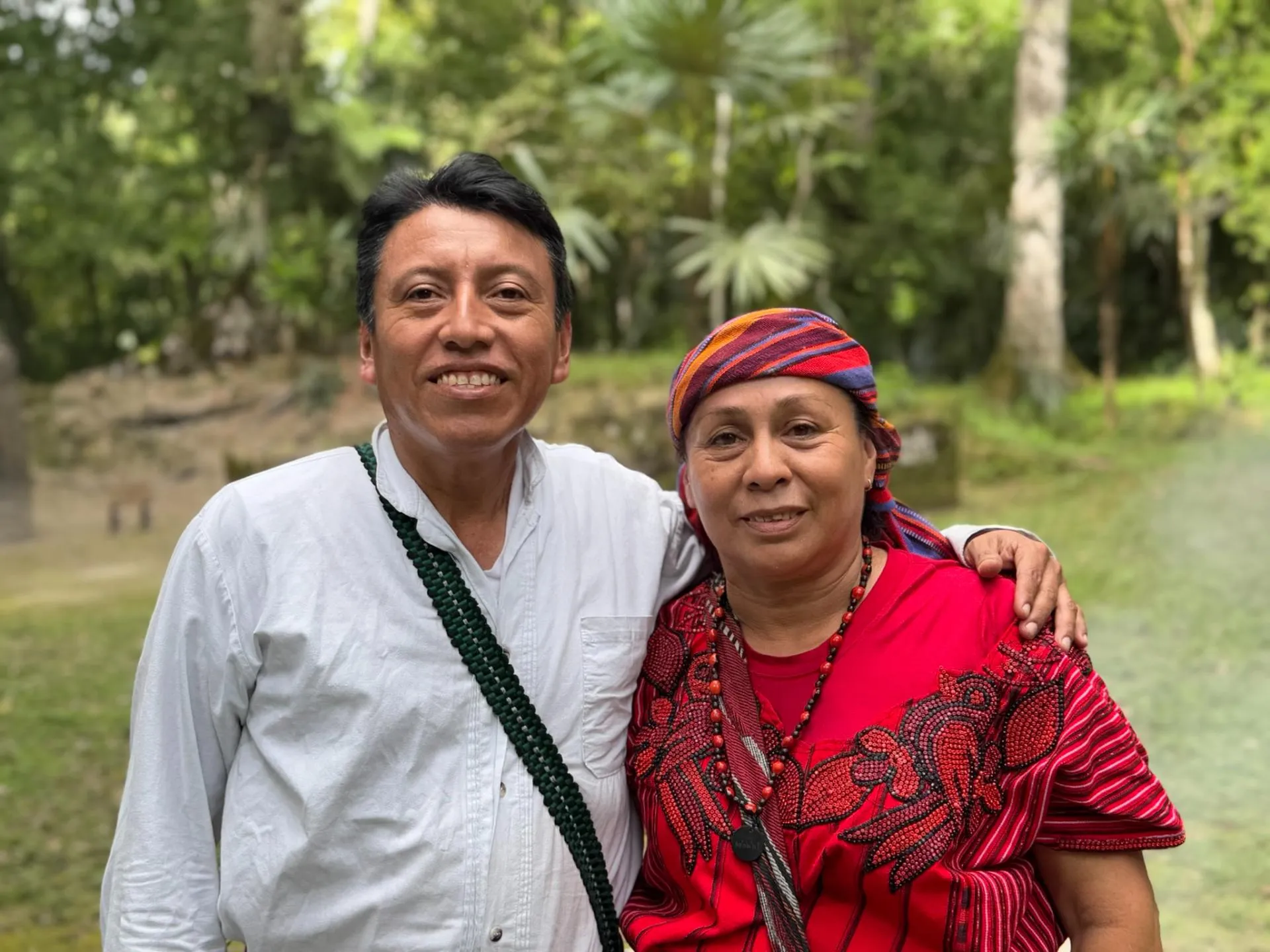 Smiling man in white shirt with a beaded necklace embracing a woman in a red traditional outfit and headscarf outdoors with greenery in the background.
