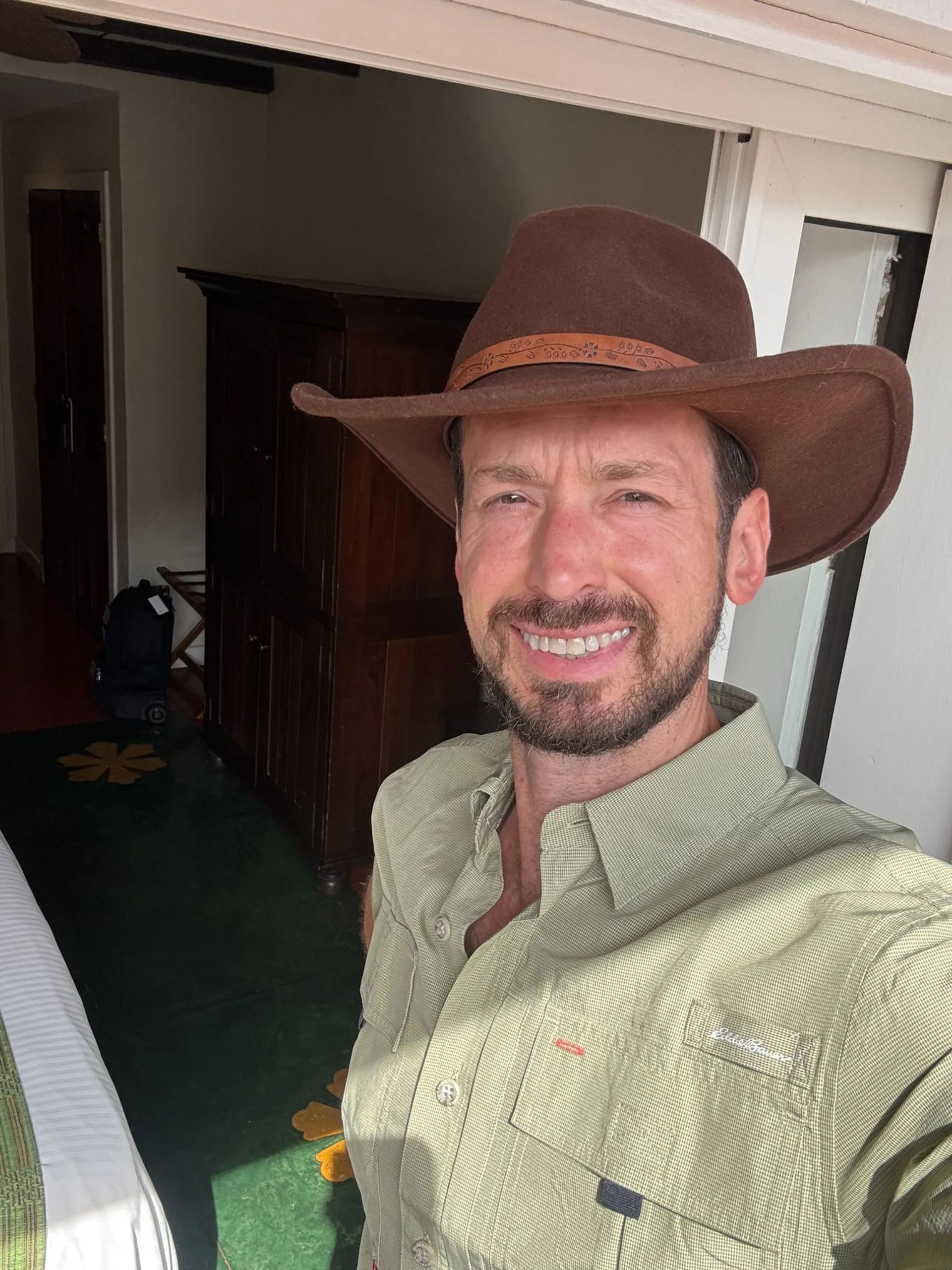 Smiling man with a beard wearing a brown wide-brimmed hat and a light green button-up shirt indoors.