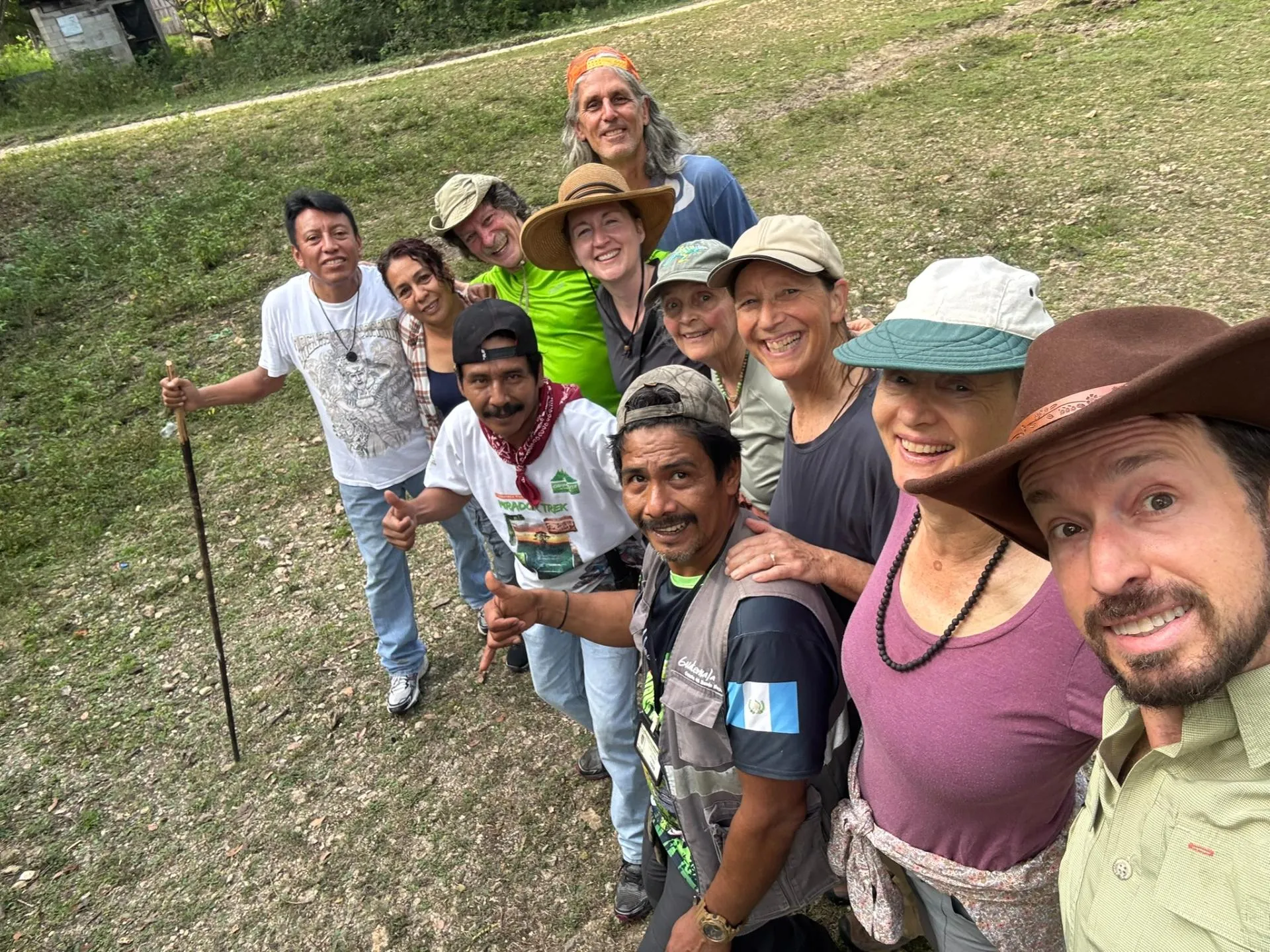 Group of people outdoors smiling at the camera, some wearing hats and casual clothing, standing on grassy ground.