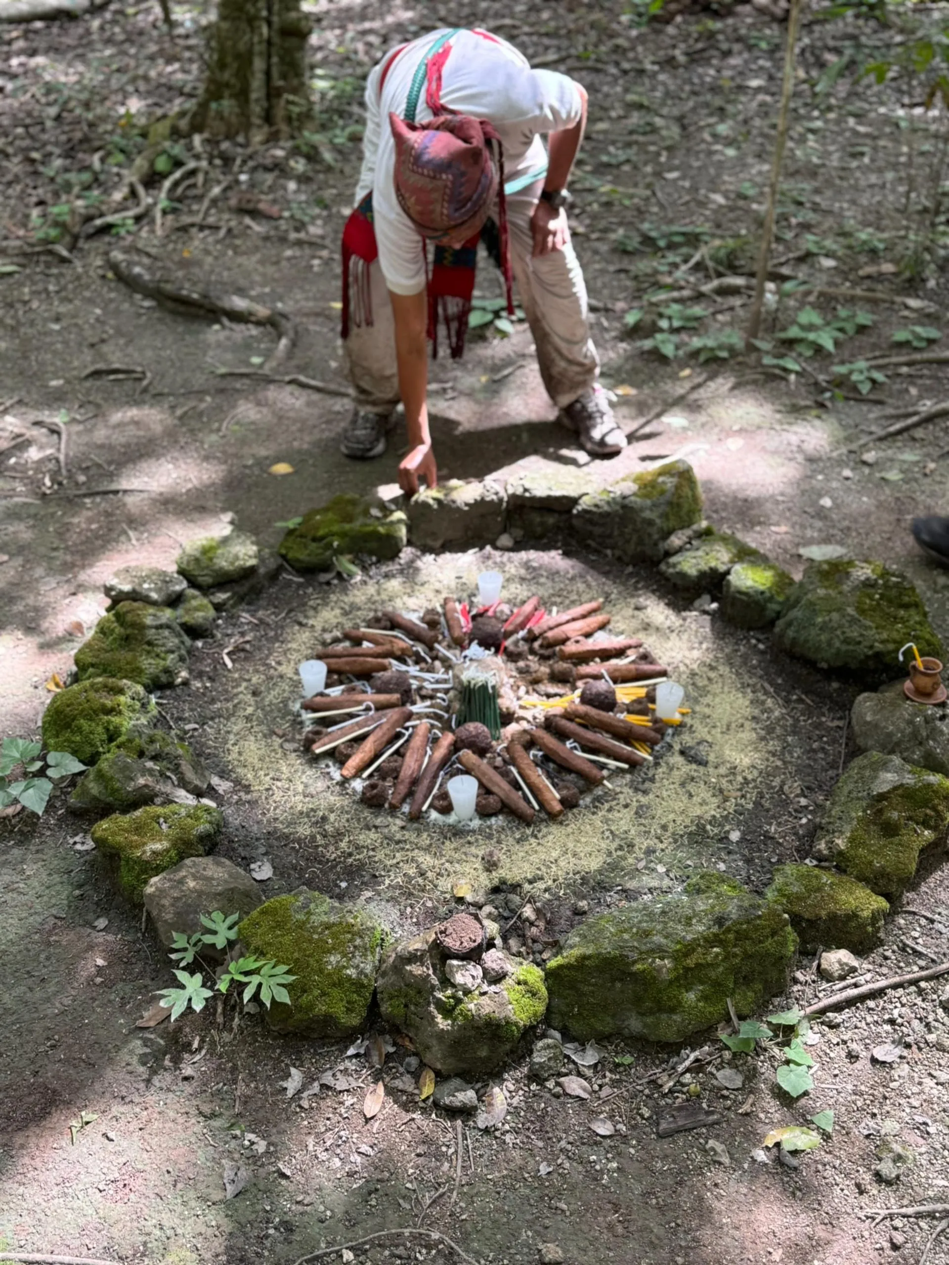 Person wearing traditional clothing places an item inside a circular stone arrangement on the forest ground containing candles and wooden sticks in a ritual setup.