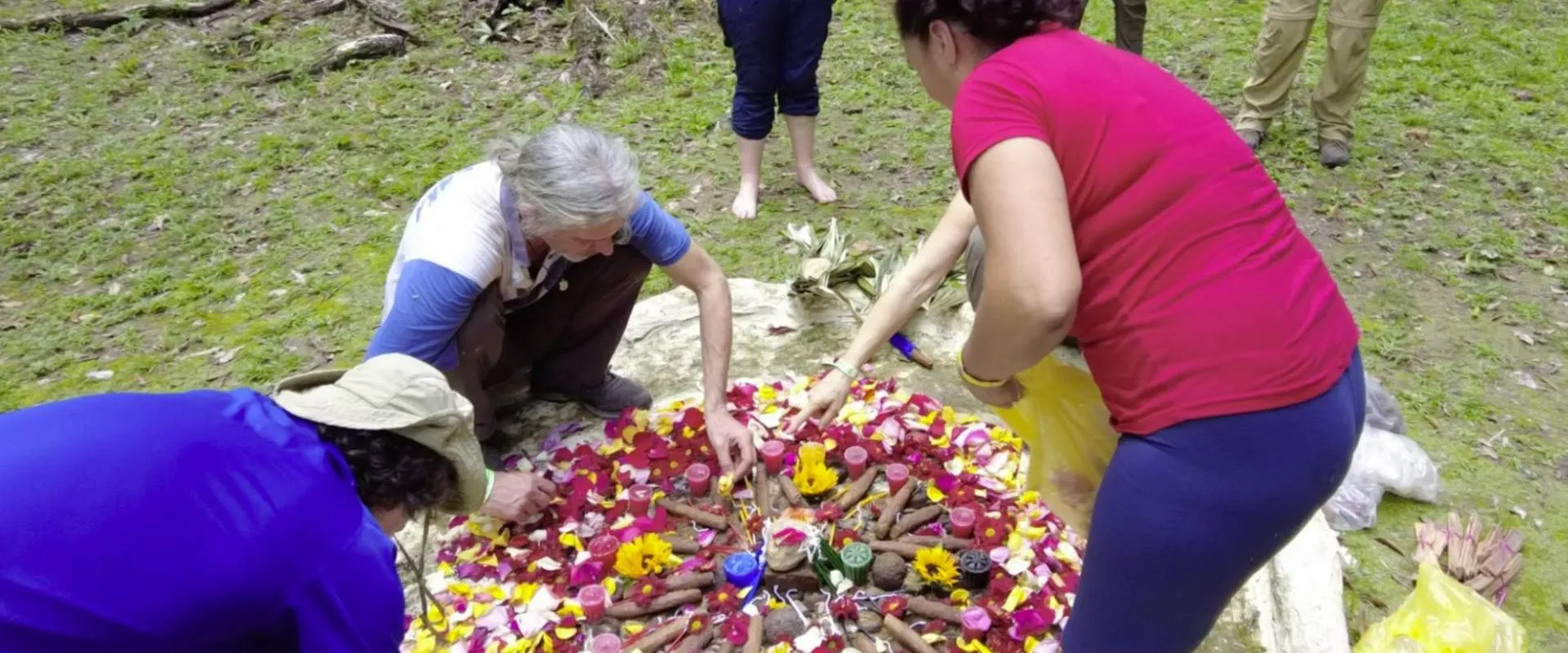 Four people outdoors arranging colorful flower petals, candles, and natural items in a circular pattern on the ground.
