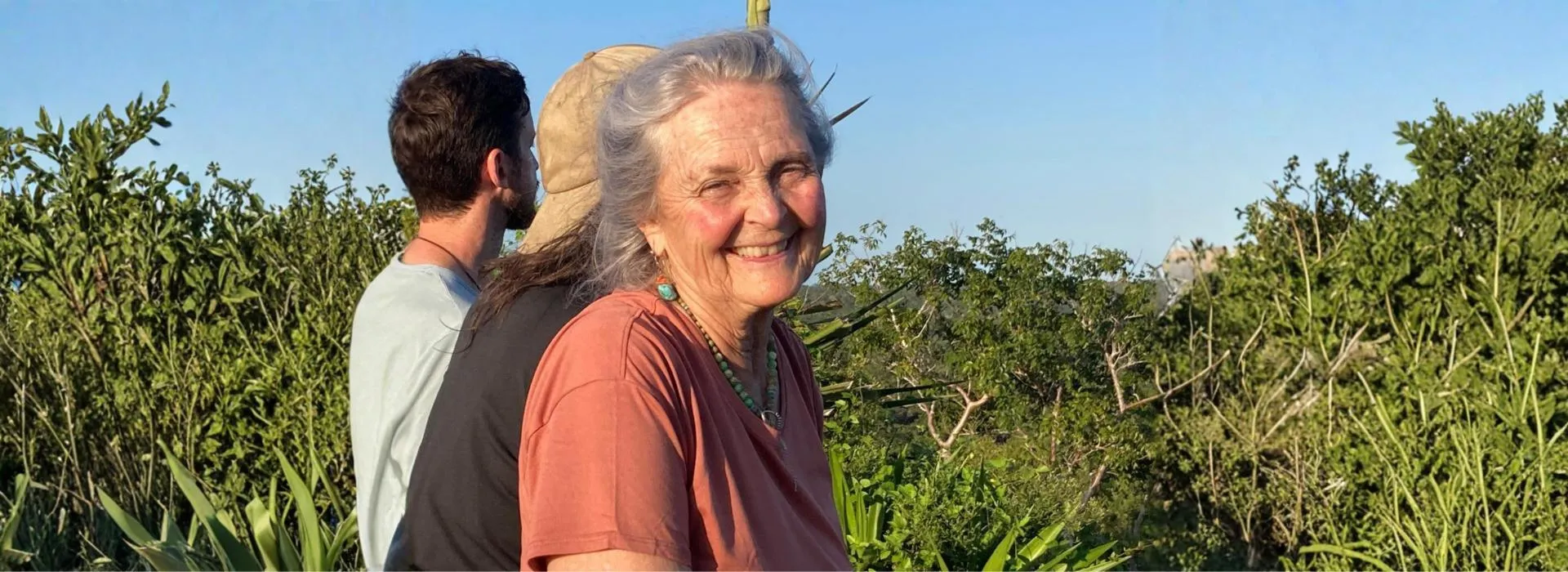 Smiling elderly woman with gray hair and turquoise jewelry outdoors in front of two people and green foliage under a clear blue sky.