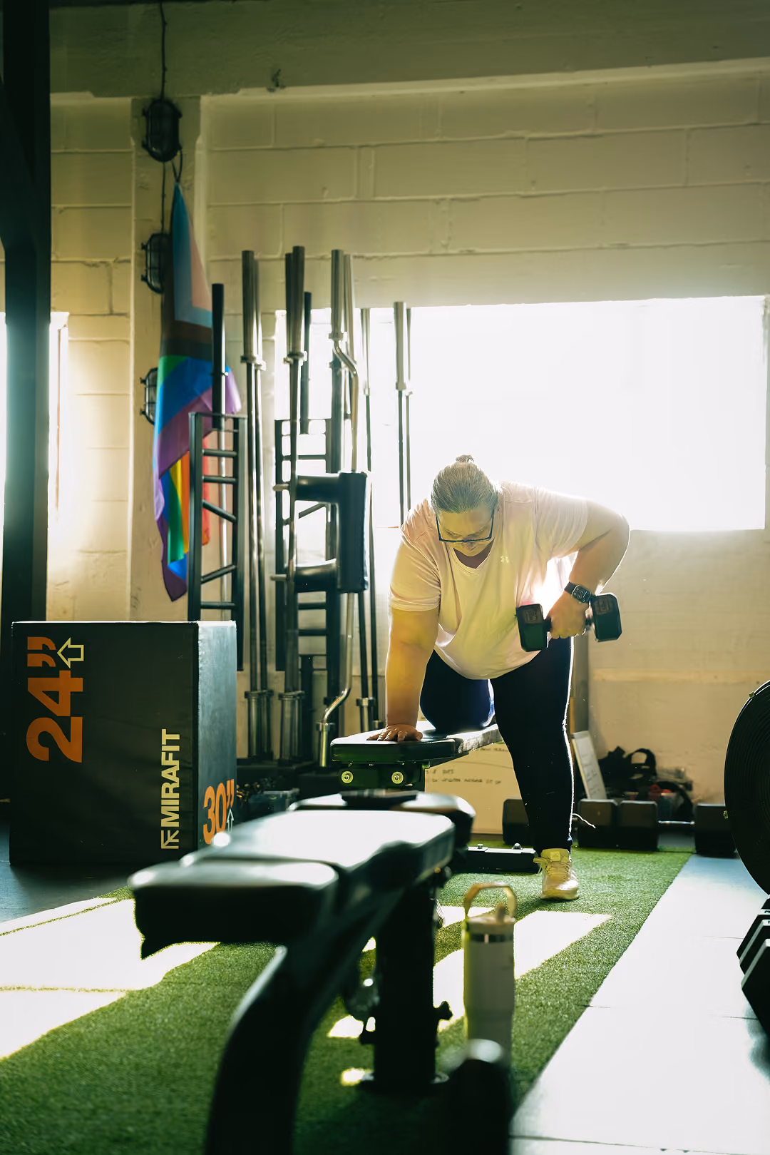 Person exercising in gym lifting dumbbell with one arm supported on bench on green turf floor.