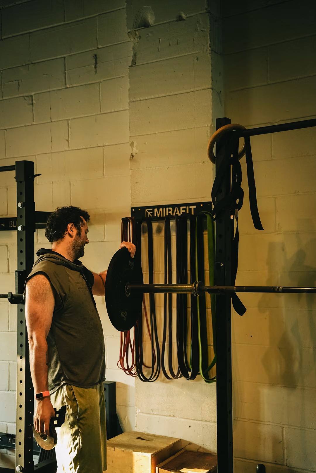 Man adjusting weight plate on barbell in gym with resistance bands hanging on wall behind him.