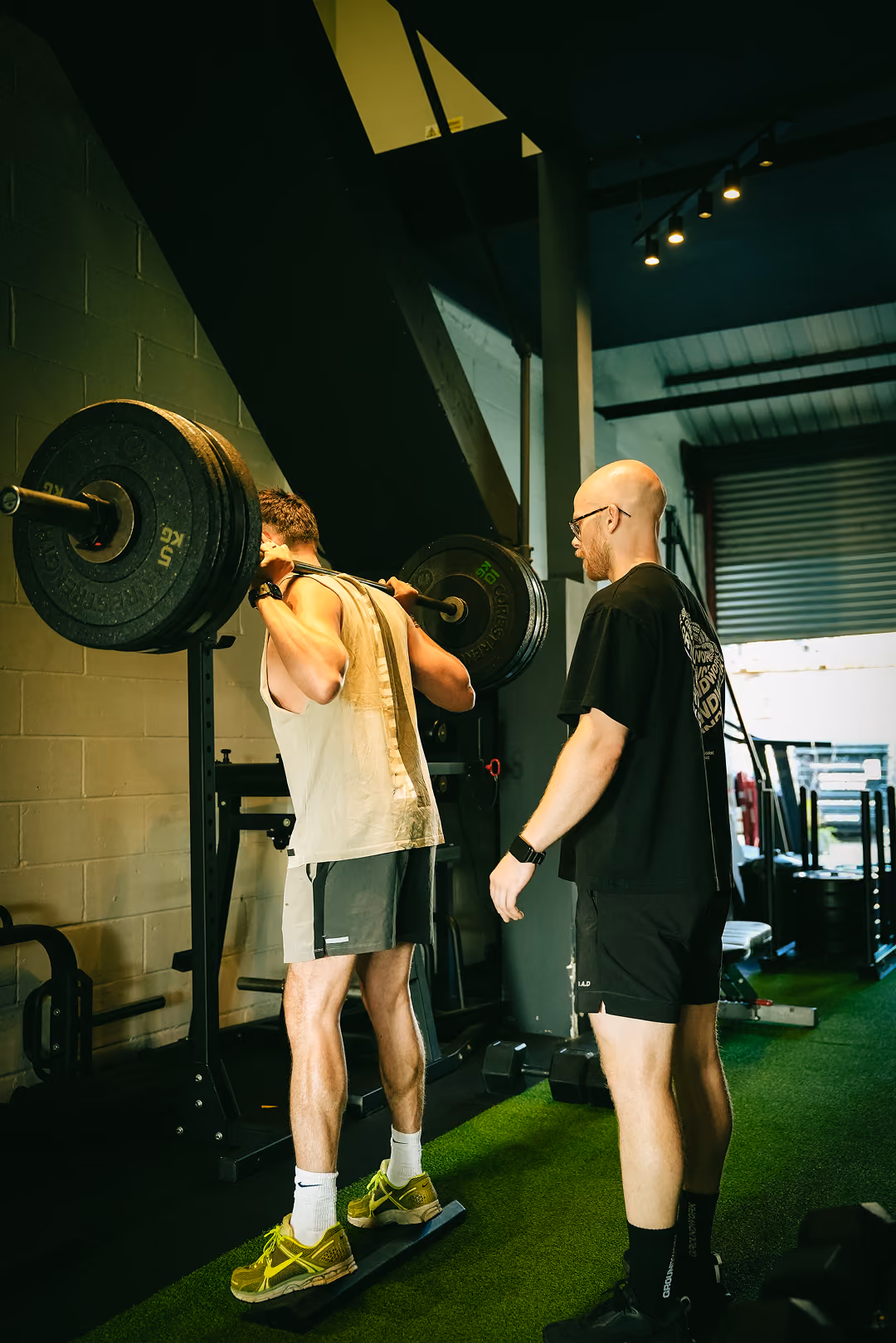 Man lifting heavy barbell on shoulders with trainer spotting him in a gym with green turf floor.