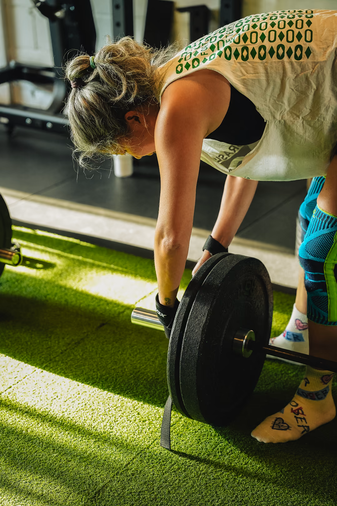 Person wearing gloves lifts a barbell with heavy weights on green turf in a gym setting.