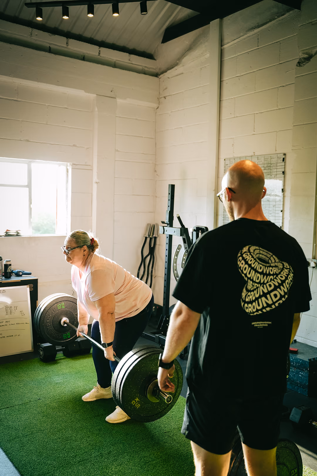 Woman in pink shirt deadlifting weights under supervision in a gym with green flooring.