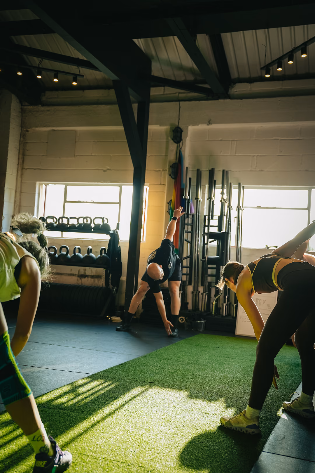 People stretching in a gym with kettlebells on a rack and weight bars against a wall.