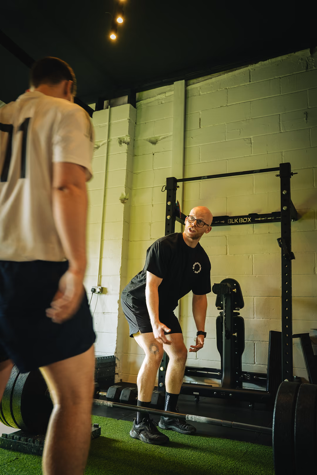 Trainer instructing a man on deadlift form near a loaded barbell in a gym with artificial turf floor.