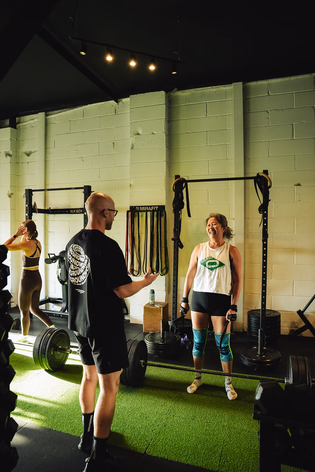 Two people smiling and talking next to a loaded barbell in a gym with green turf flooring.