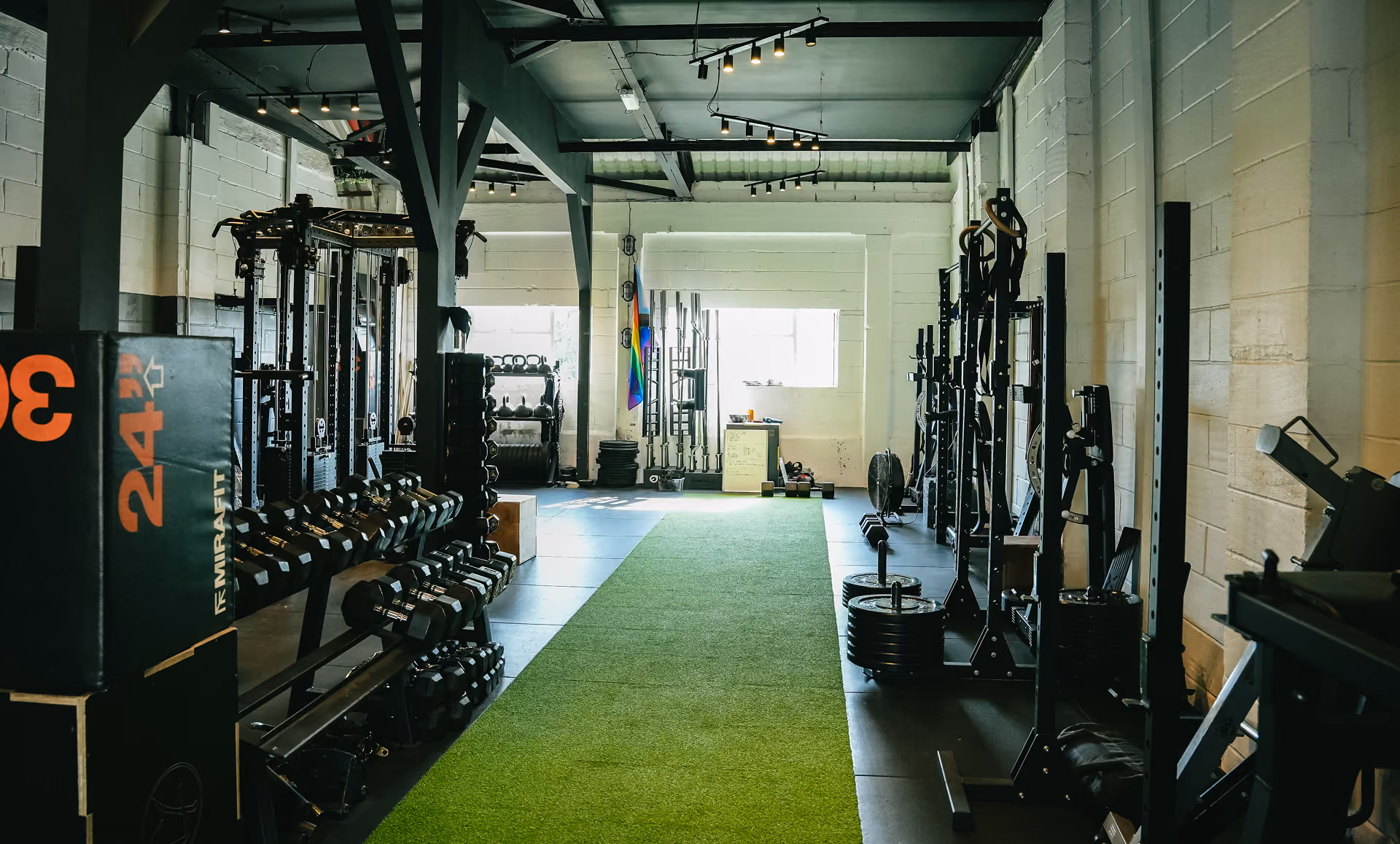 Interior view of a gym with dumbbells, weights, racks, and a green turf running down the center.