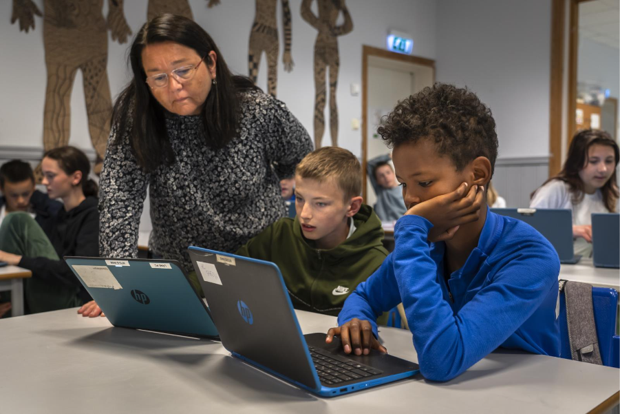 To elever som løser matteoppgaver paa hver sin pc. En lærer staar bøyd over pcen til den ene eleven. Alle sitter og tenker.