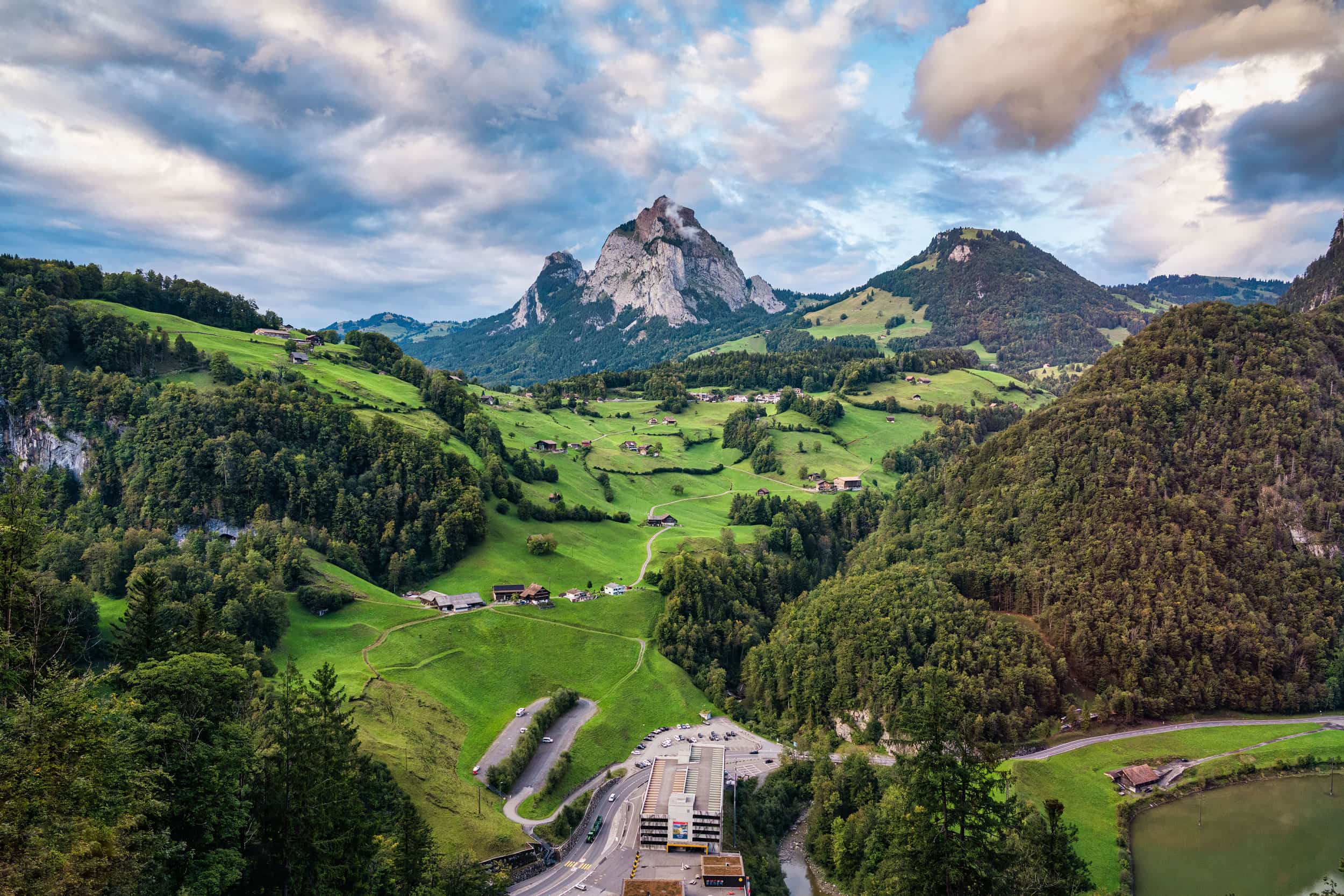 Green alpine valley with scattered chalets, forested hills and rocky mountain peaks in Switzerland.
