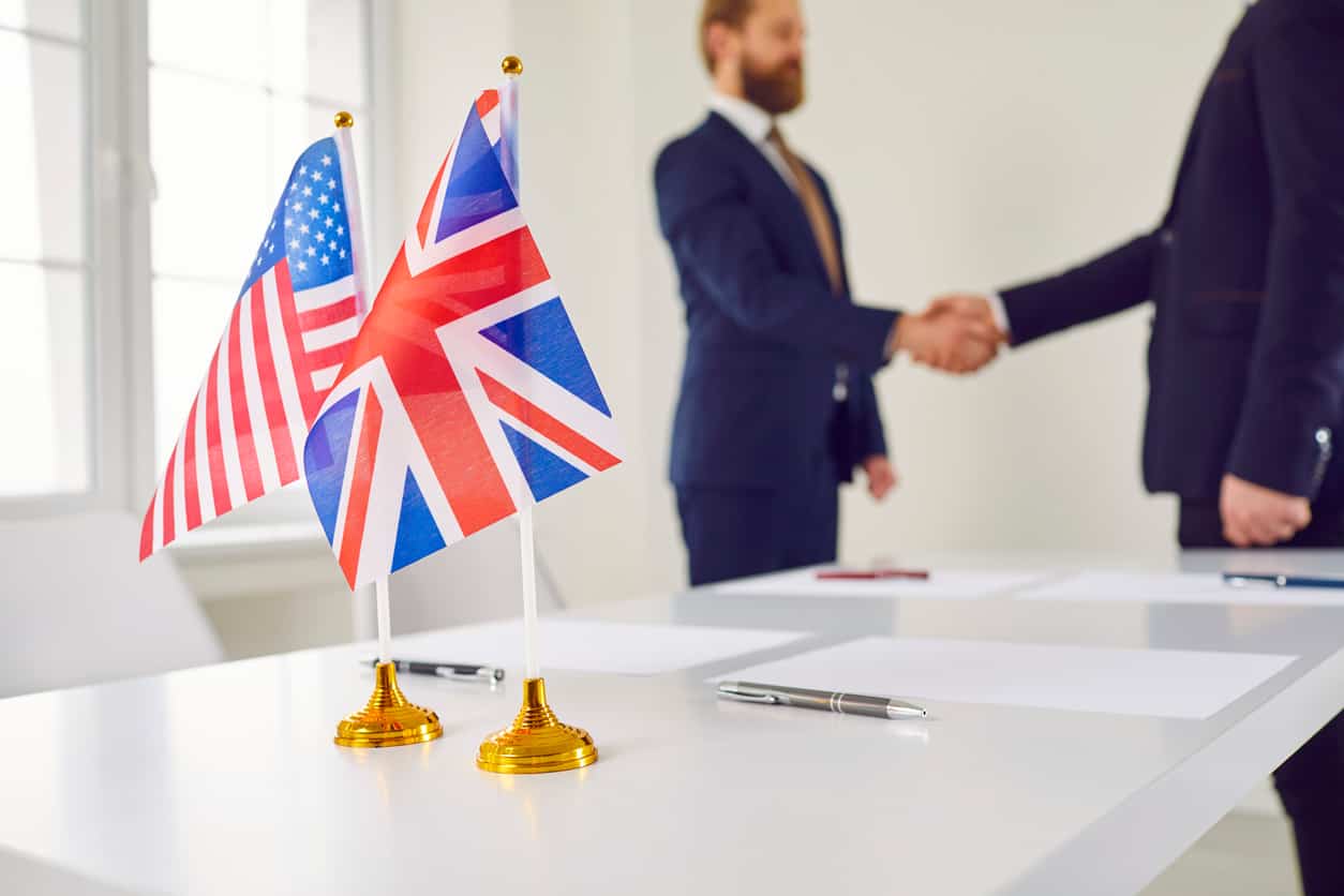United States and United Kingdom flags with businesspeople shaking hands in an office