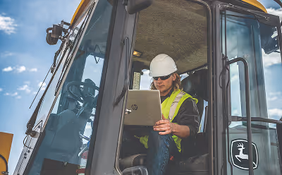 Construction worker wearing a white hard hat and yellow safety vest using a laptop inside a vehicle cabin.