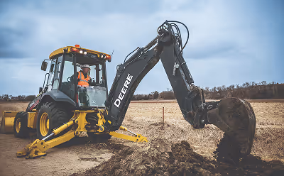 Yellow and black John Deere excavator digging soil in a field with an operator inside the cab.