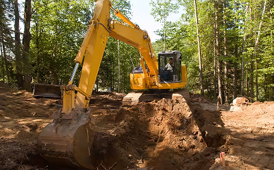 Yellow excavator digging soil in a forested area with trees around.